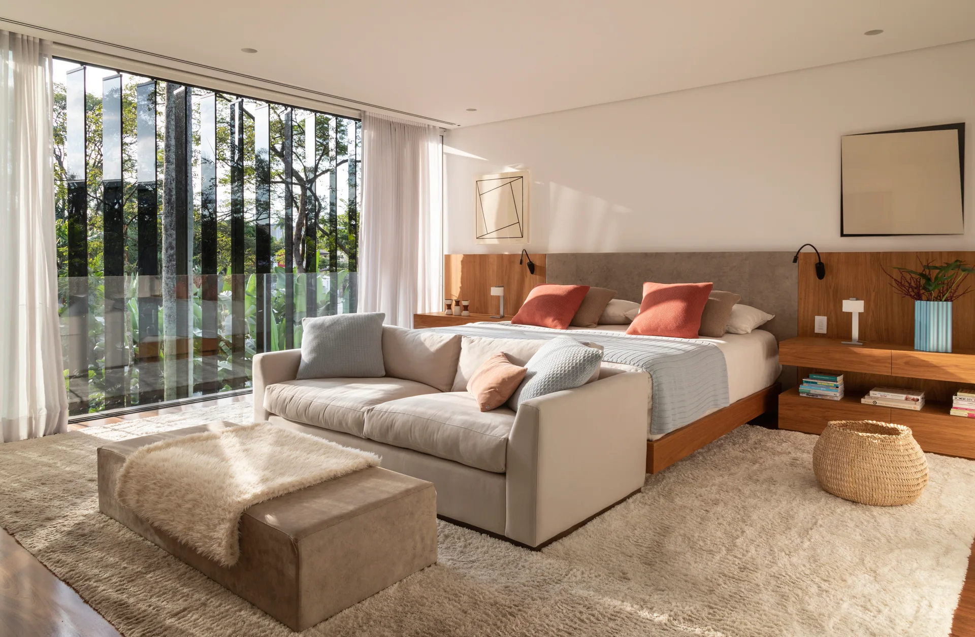A contemporary bedroom featuring an integrated wood headboard wall, a cream sofa at the foot of the bed, and a large glass window looking out at trees.