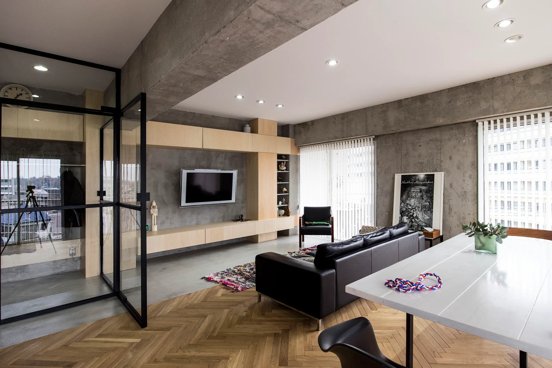 A modern living space featuring a black leather sofa, raw concrete walls, custom light wood media built-ins, and herringbone patterned wood floors.