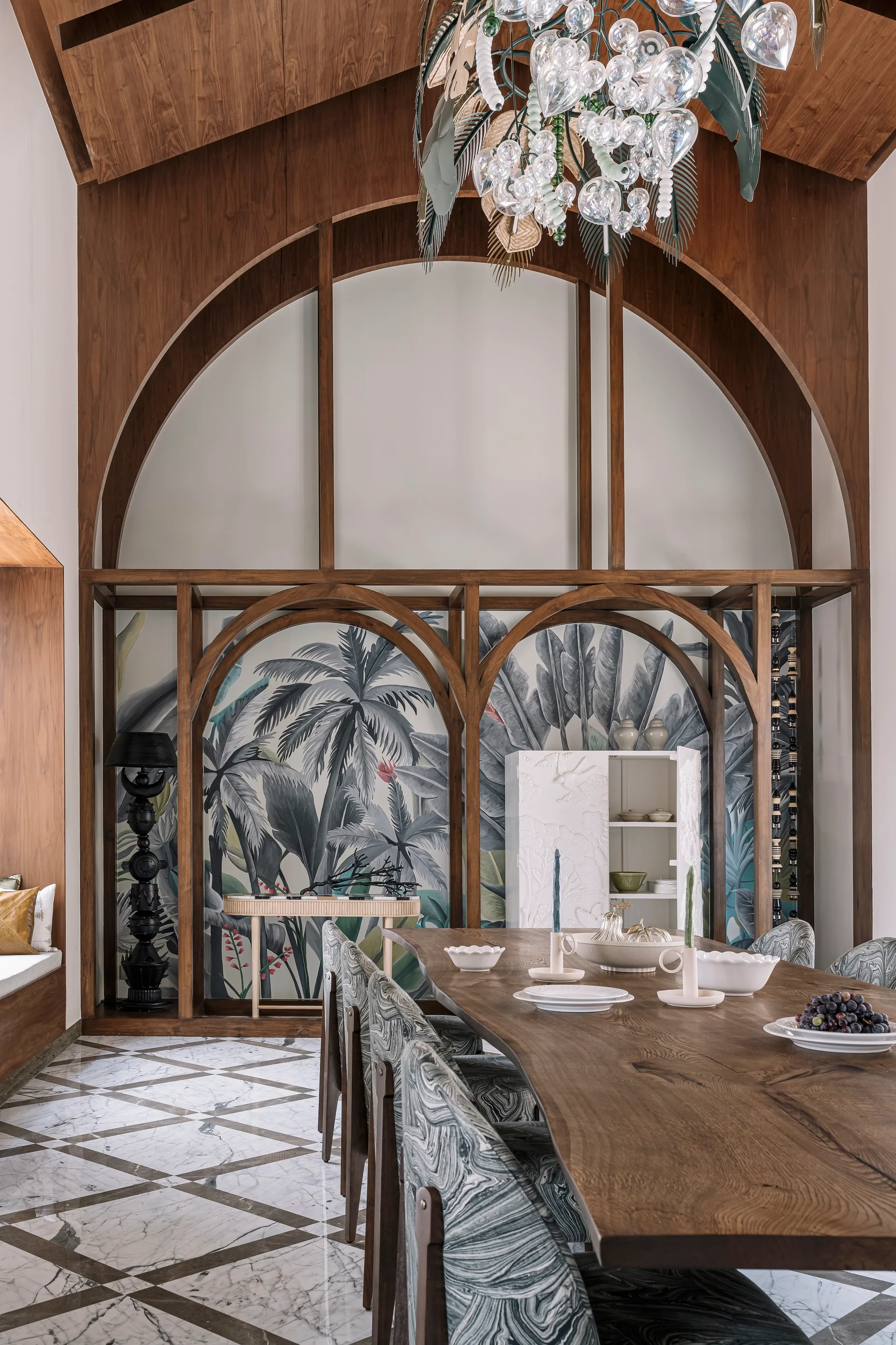 A contemporary dining room features a long wooden table, patterned chairs, and a large wooden arch partition over tropical wallpaper.