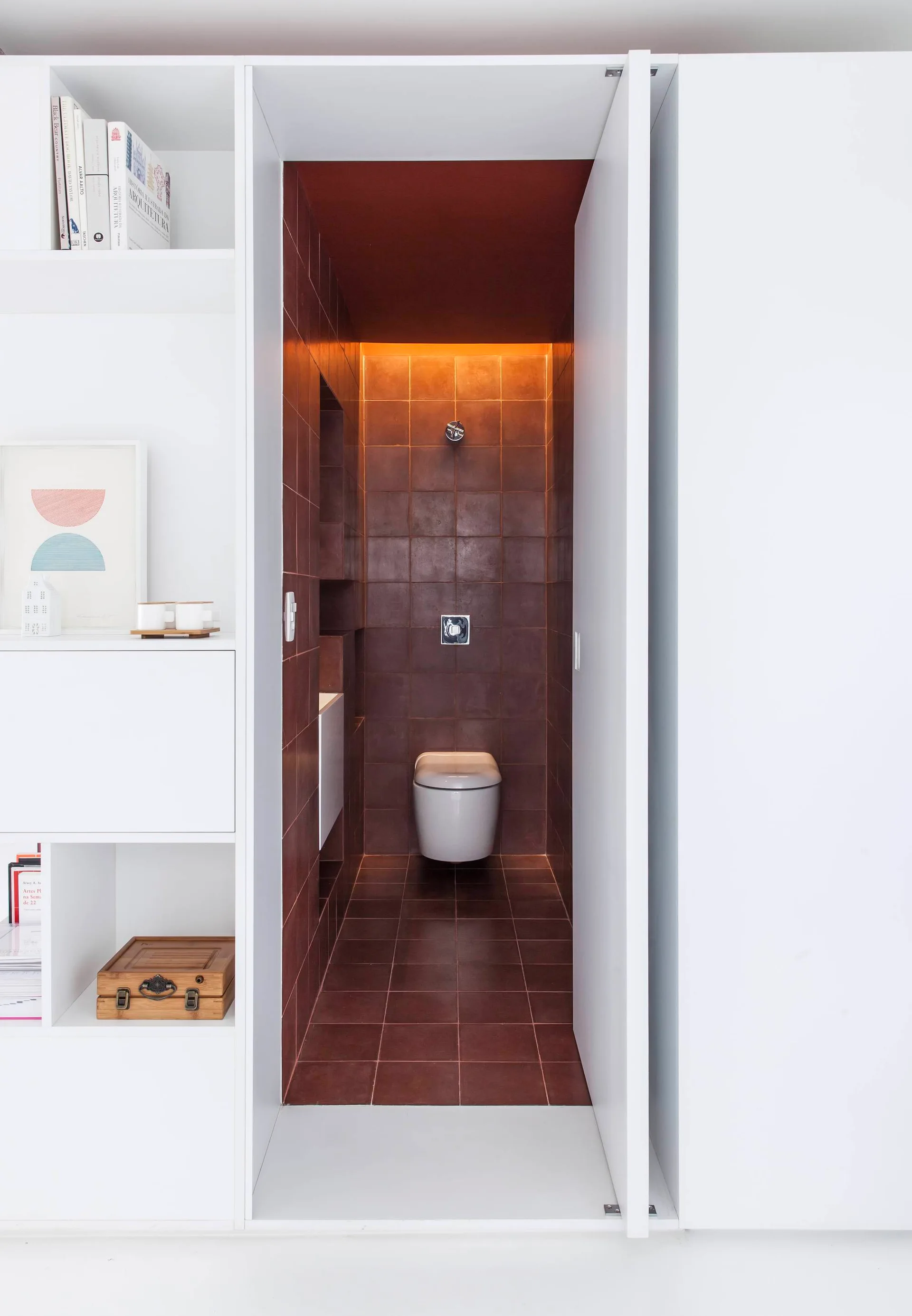 A minimalist powder room lined with dark red square tiles is framed by a white built-in bookcase holding books and art.