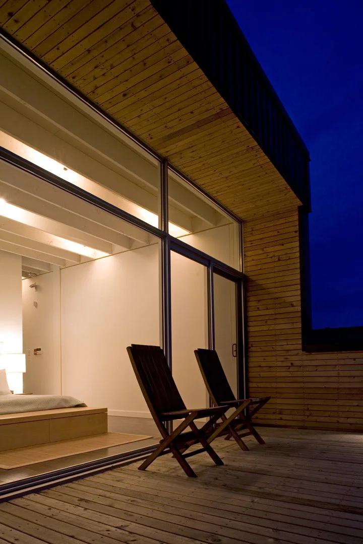 Two wooden folding chairs sit on a timber deck outside a bedroom with large glass sliding doors and warm interior lighting at night.