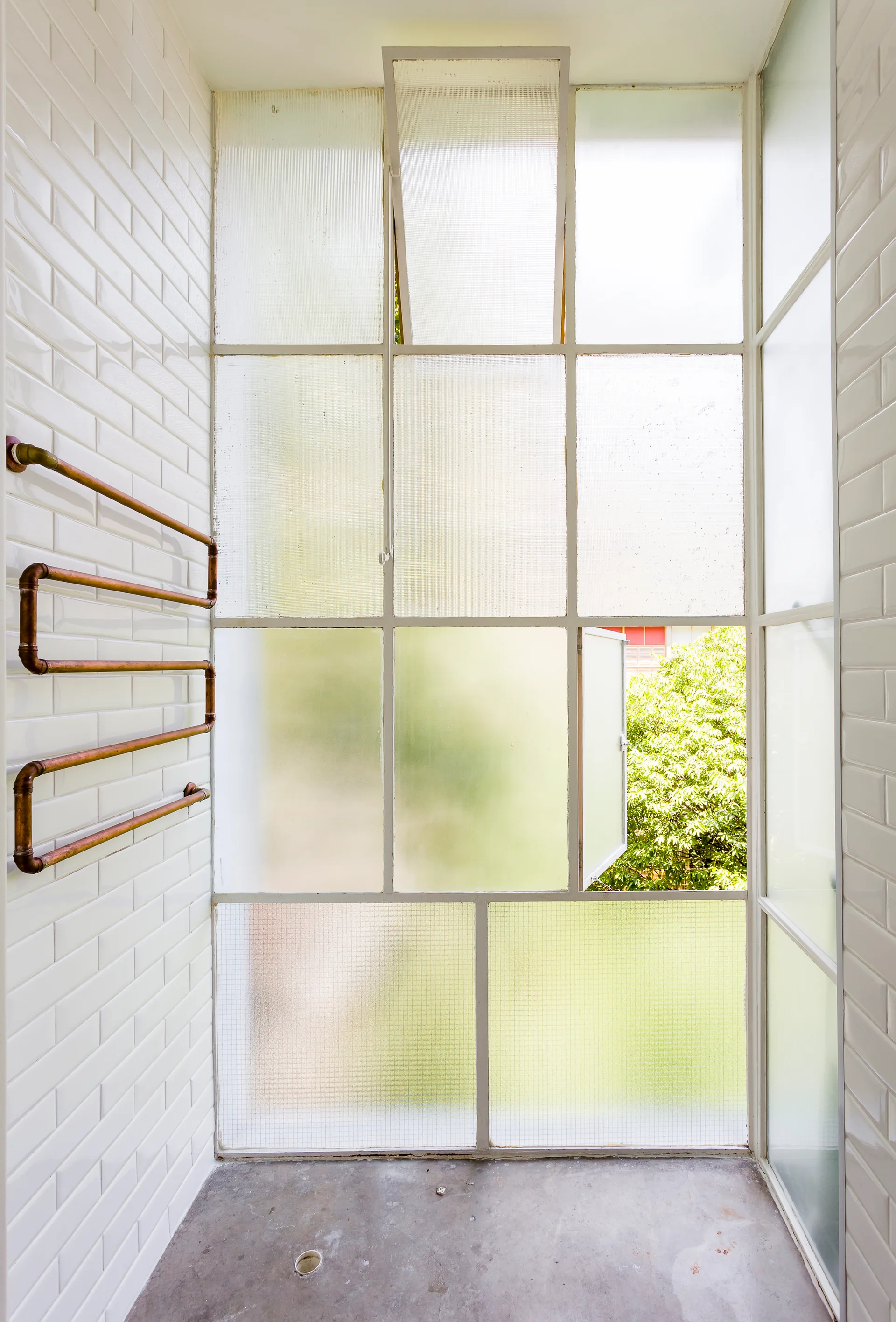 A bright bathroom interior featuring white subway tiled walls, a large gridded frosted glass window, and a wall-mounted copper pipe towel warmer.
