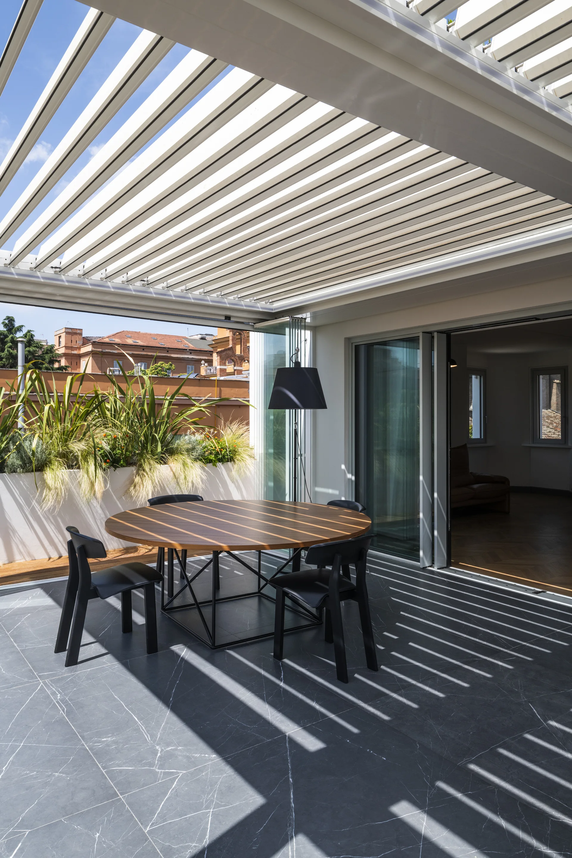 A round wood-topped dining table with black chairs sits on a dark gray tiled terrace under a white louvered pergola casting linear shadows.