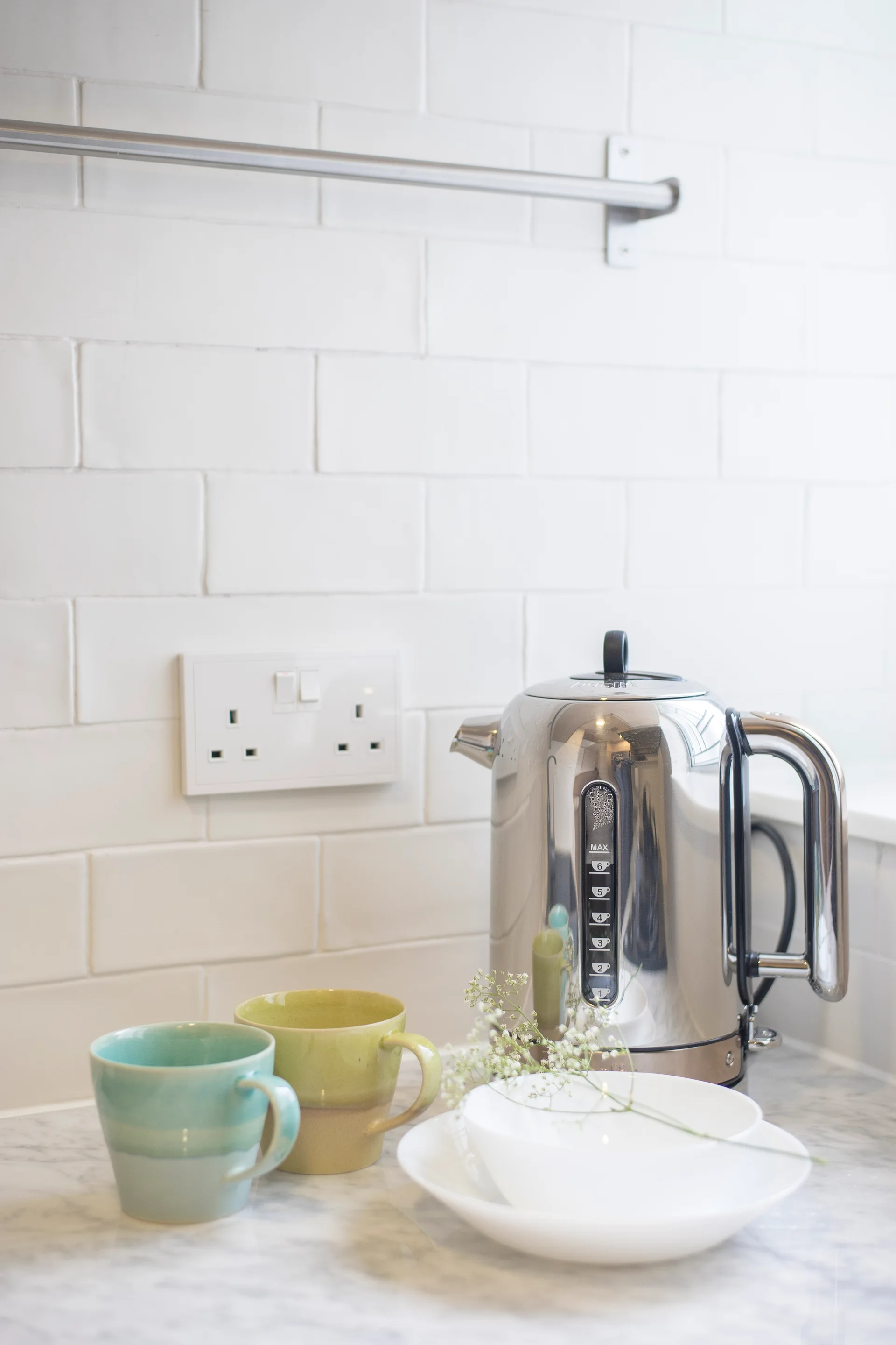 A chrome electric kettle sits next to two ceramic mugs on a marble kitchen countertop in front of a white subway tile backsplash.