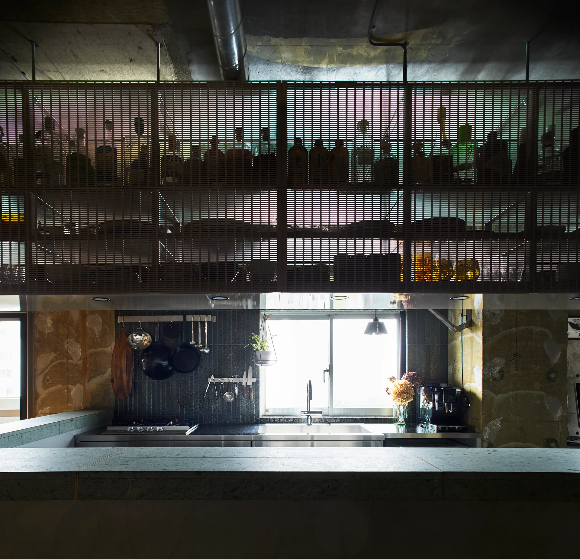 An industrial kitchen with a raw concrete ceiling, a dark tiled backsplash, and suspended metal mesh storage above the counter.