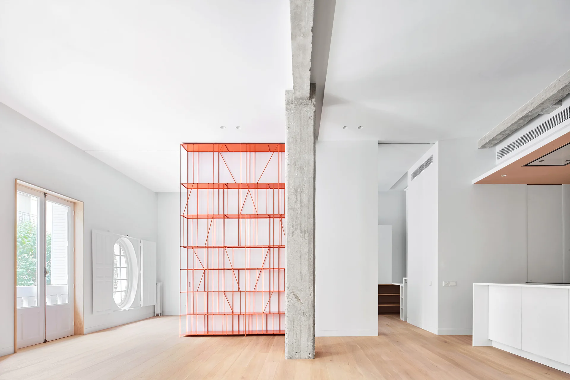 A minimalist white open-plan living room featuring a tall orange metal shelving unit, a raw concrete column, and light wood flooring.