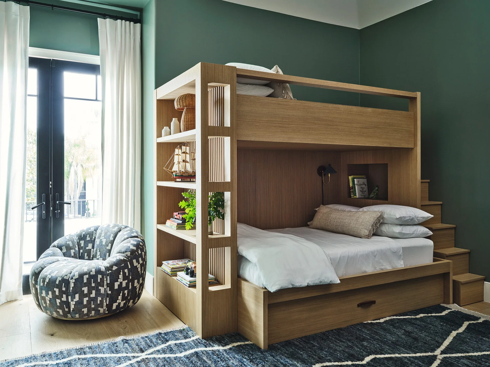 A custom wooden bunk bed with integrated shelving and stairs stands against a deep green wall next to a patterned lounge chair.