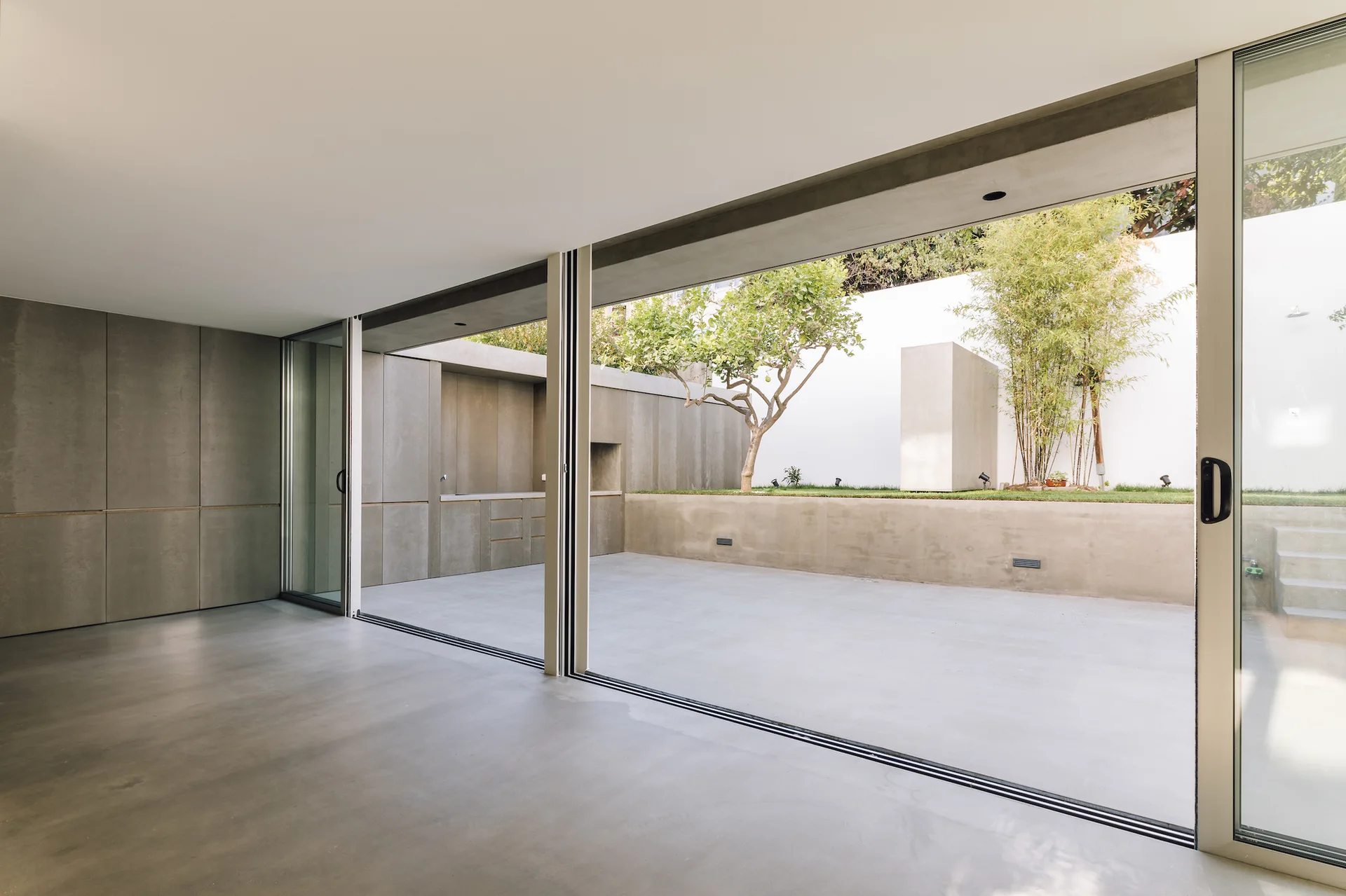An empty minimalist interior with smooth concrete floors opening through large sliding glass doors to an outdoor concrete patio.