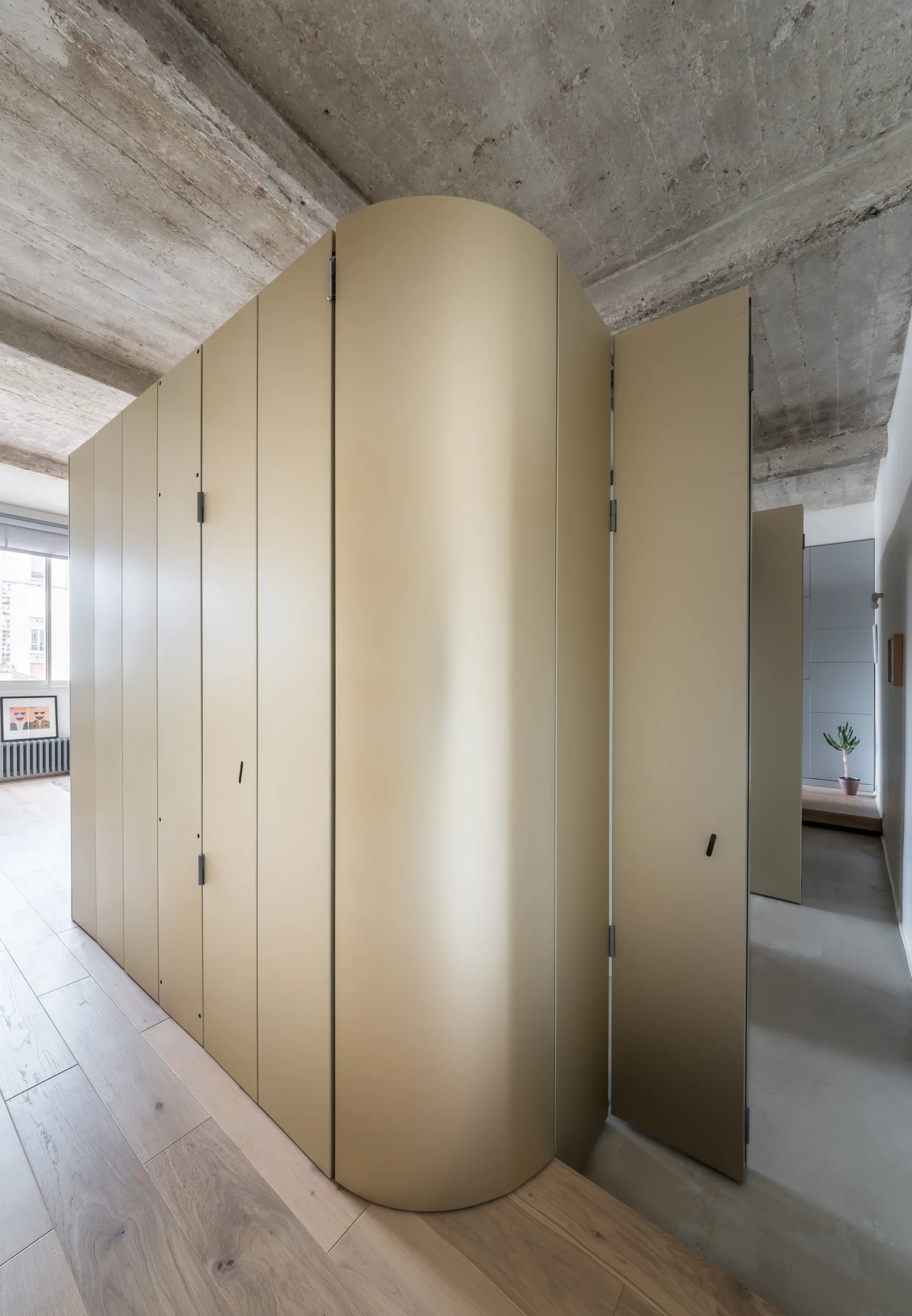 A large floor-to-ceiling room divider cabinet with a matte gold finish and a curved corner stands under an exposed concrete ceiling on wood floors.