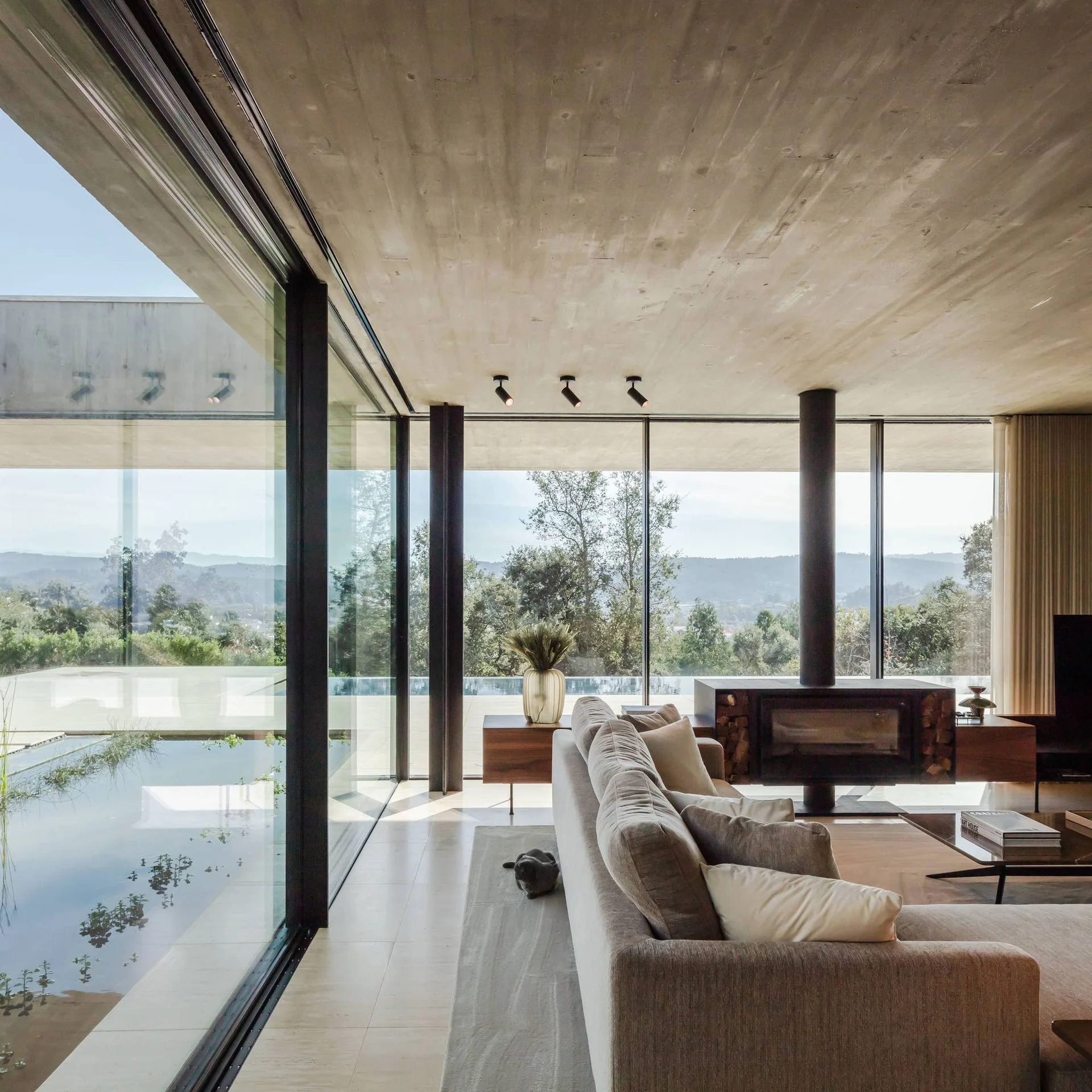 A minimalist living room features a textured concrete ceiling, a low beige sofa, a black fireplace, and floor-to-ceiling glass windows.