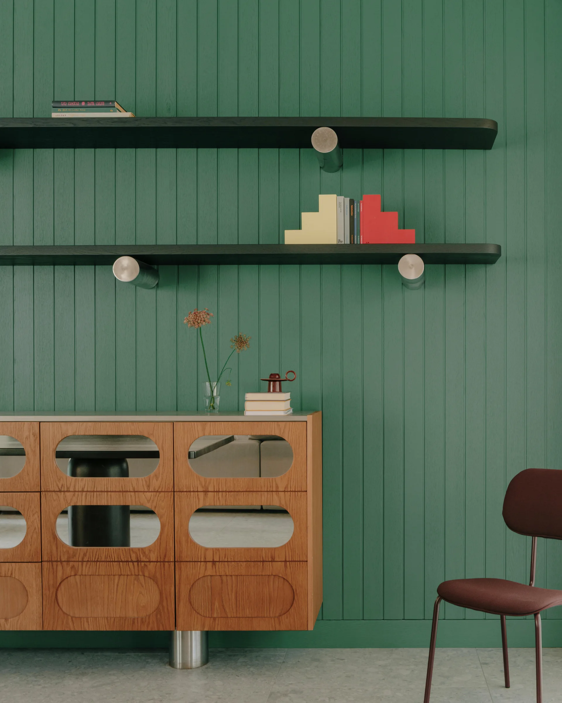 A wooden sideboard with oval mirrored inserts stands against a green vertically paneled wall below two black floating shelves.