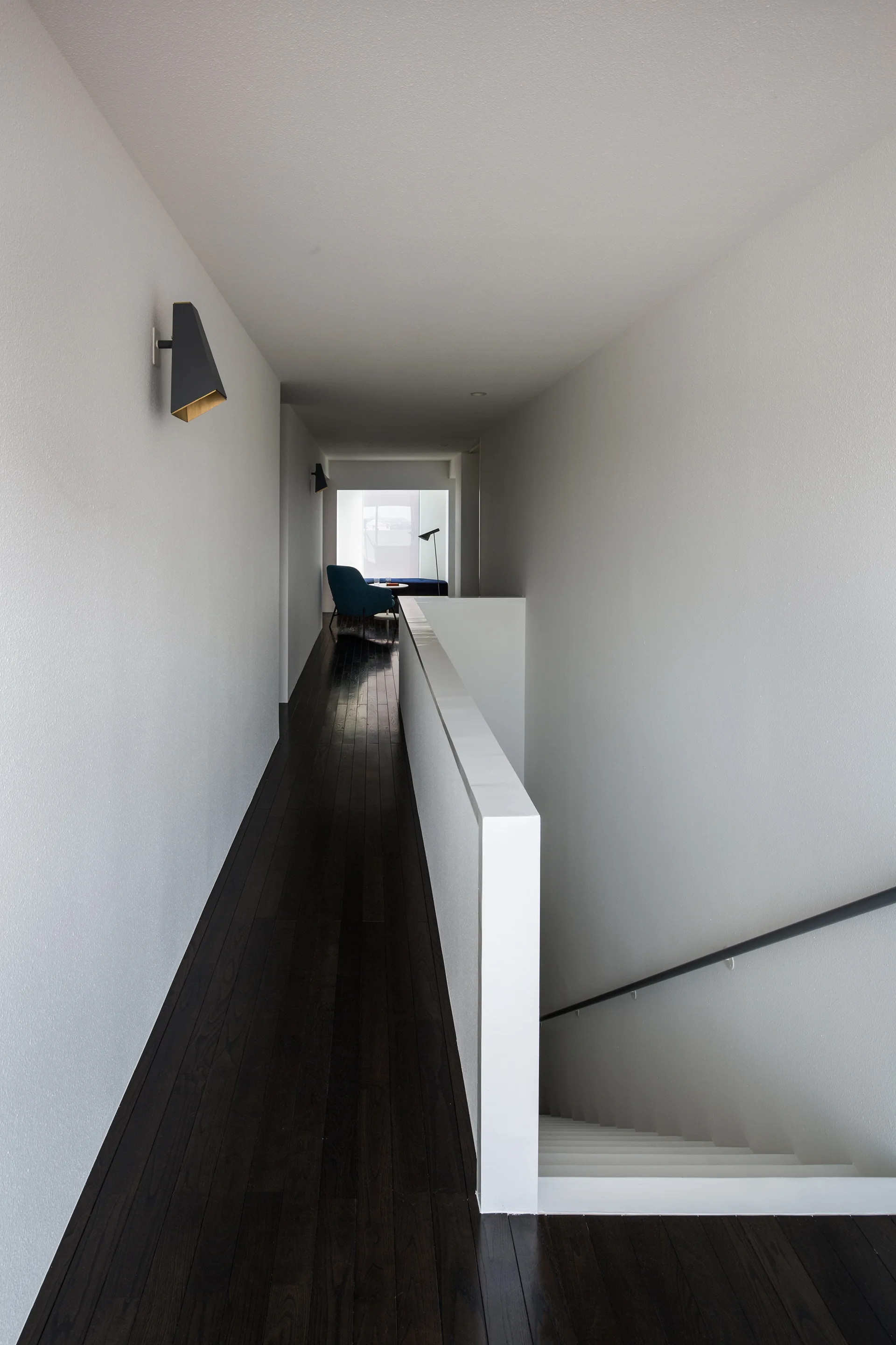 A narrow white hallway features dark wood flooring, a solid white stairwell guardrail, and geometric black wall sconces.
