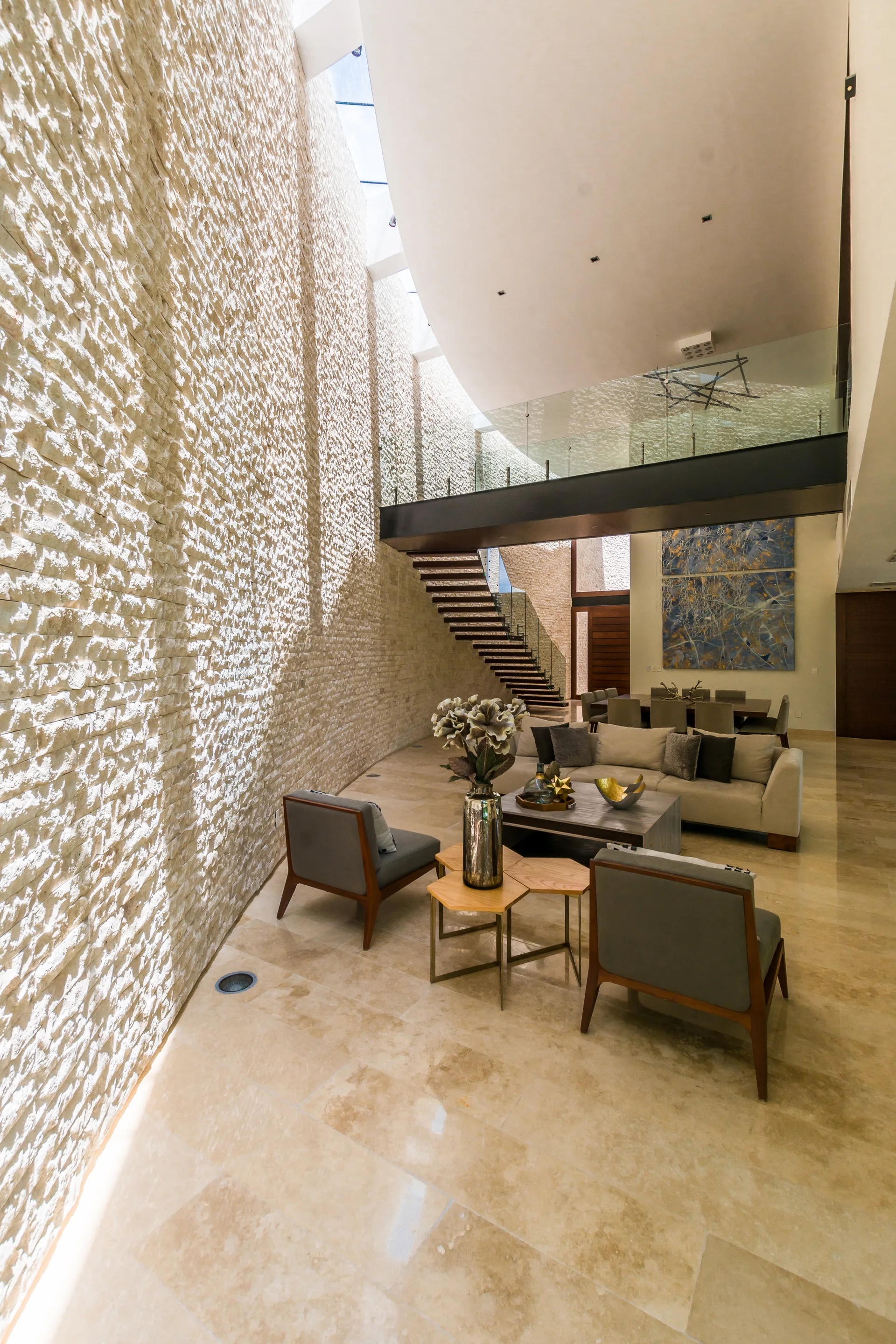 A spacious living room featuring a tall, textured stone wall, a floating wooden staircase, polished stone floor tiles, and neutral furniture.