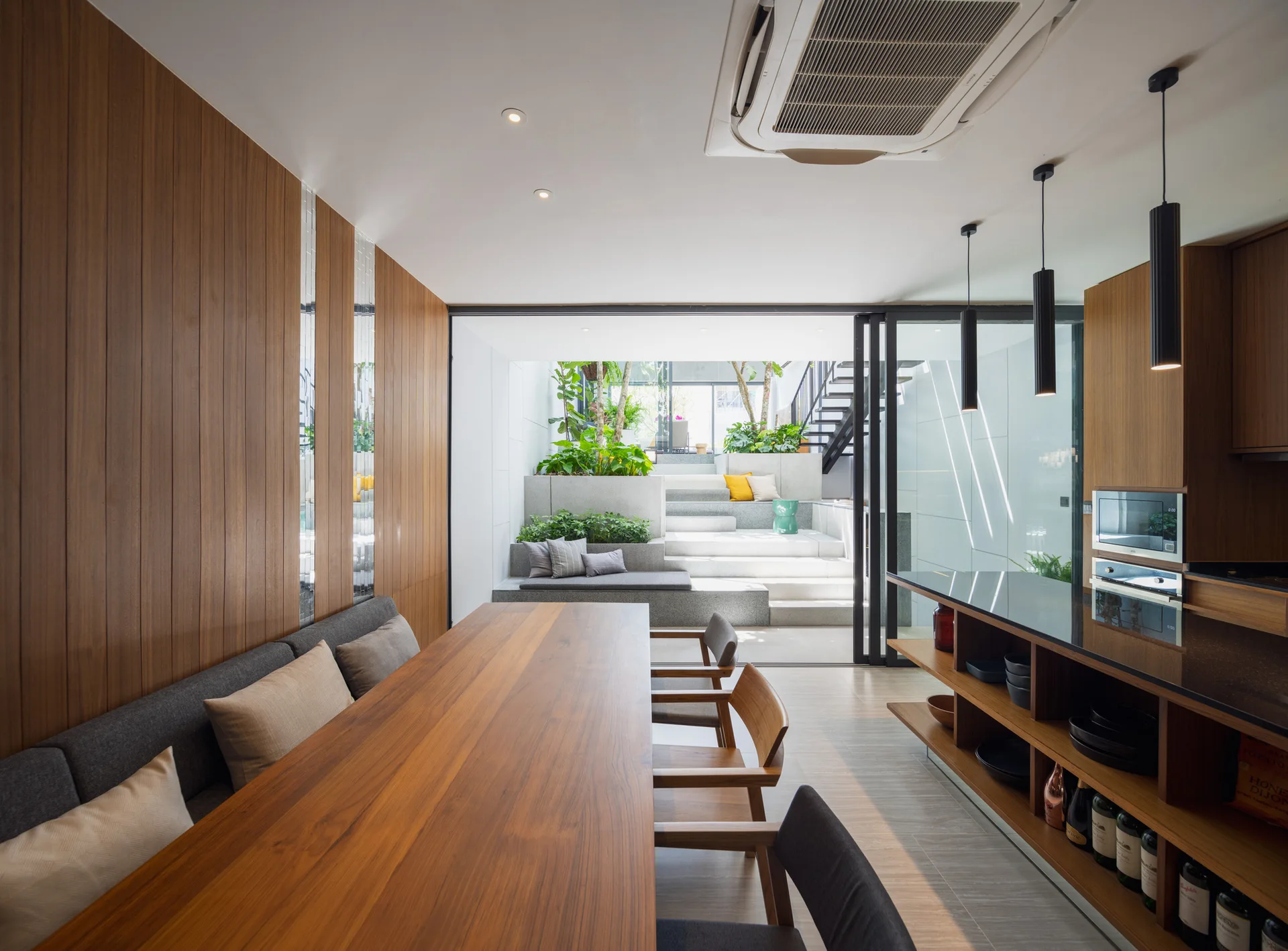 A dining room features a long wooden table, built-in banquette, vertical wood wall paneling, and glass sliding doors opening to an outdoor courtyard.