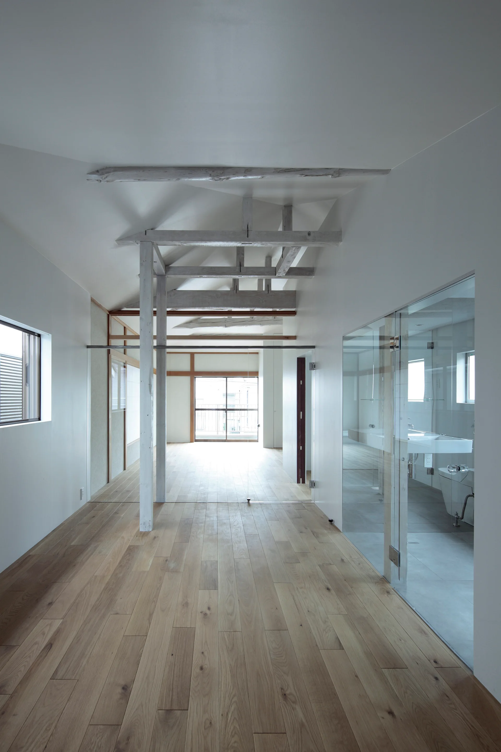A minimalist hallway features light hardwood flooring, white vaulted ceilings with exposed timber beams, and a glass-enclosed bathroom.