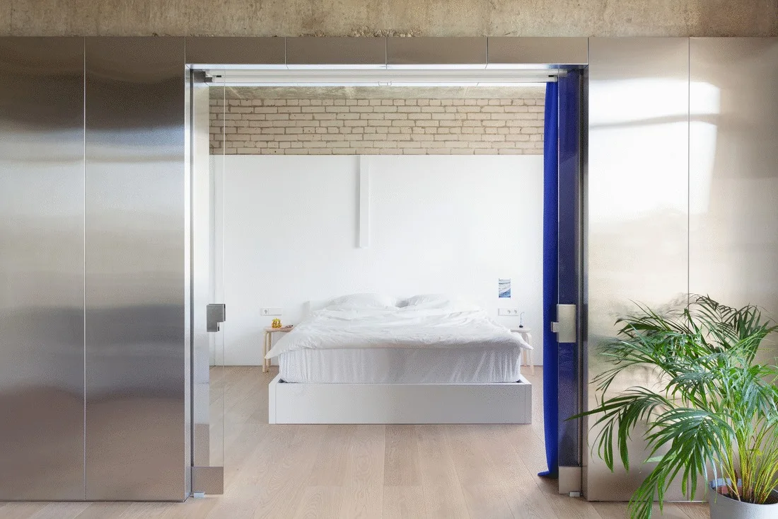 A minimalist bedroom featuring a white bed framed by reflective stainless steel partitions and an exposed brick accent wall.
