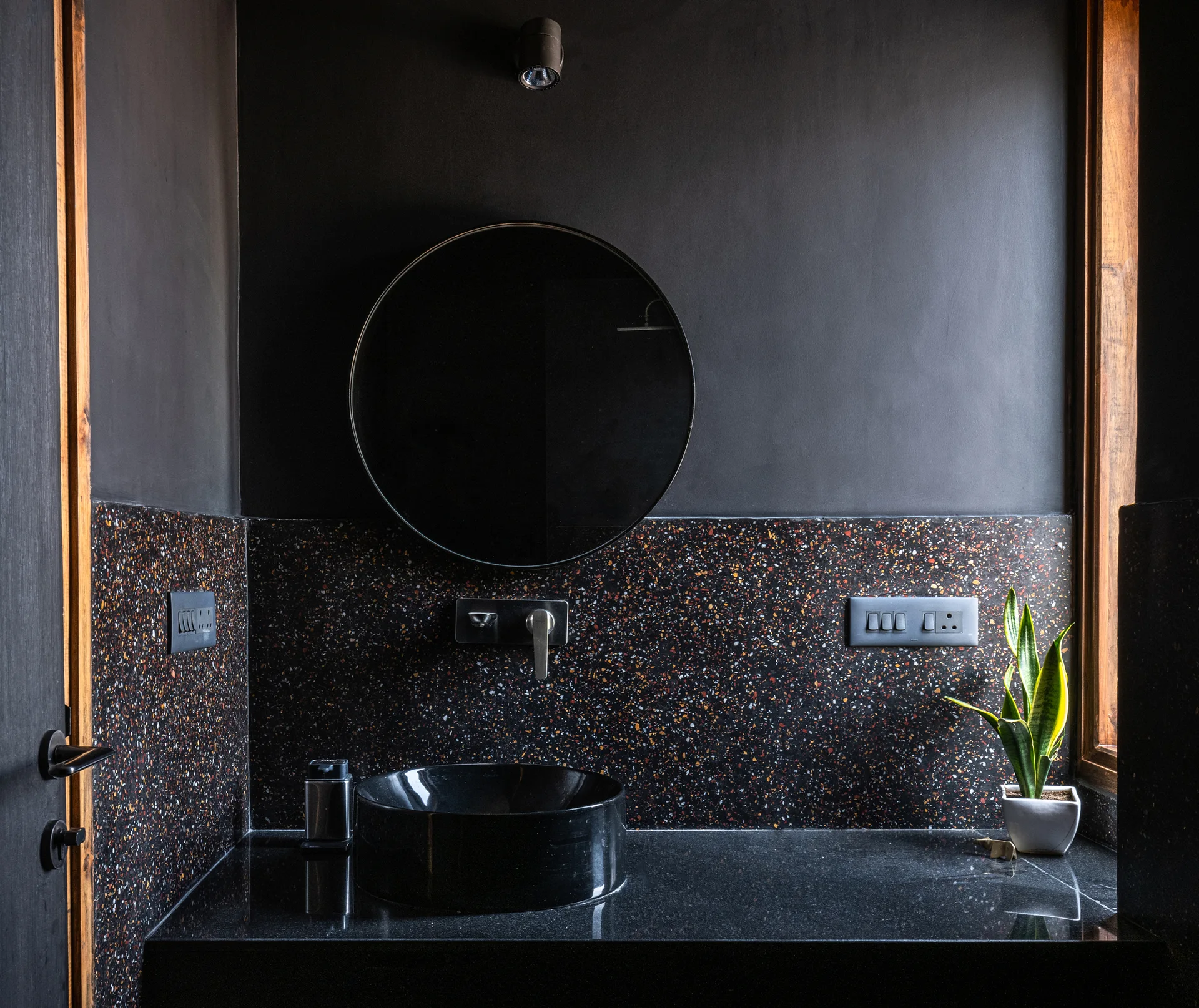 A dark bathroom vanity features a black vessel sink, a round mirror on a matte black wall, and a colorful speckled terrazzo backsplash.