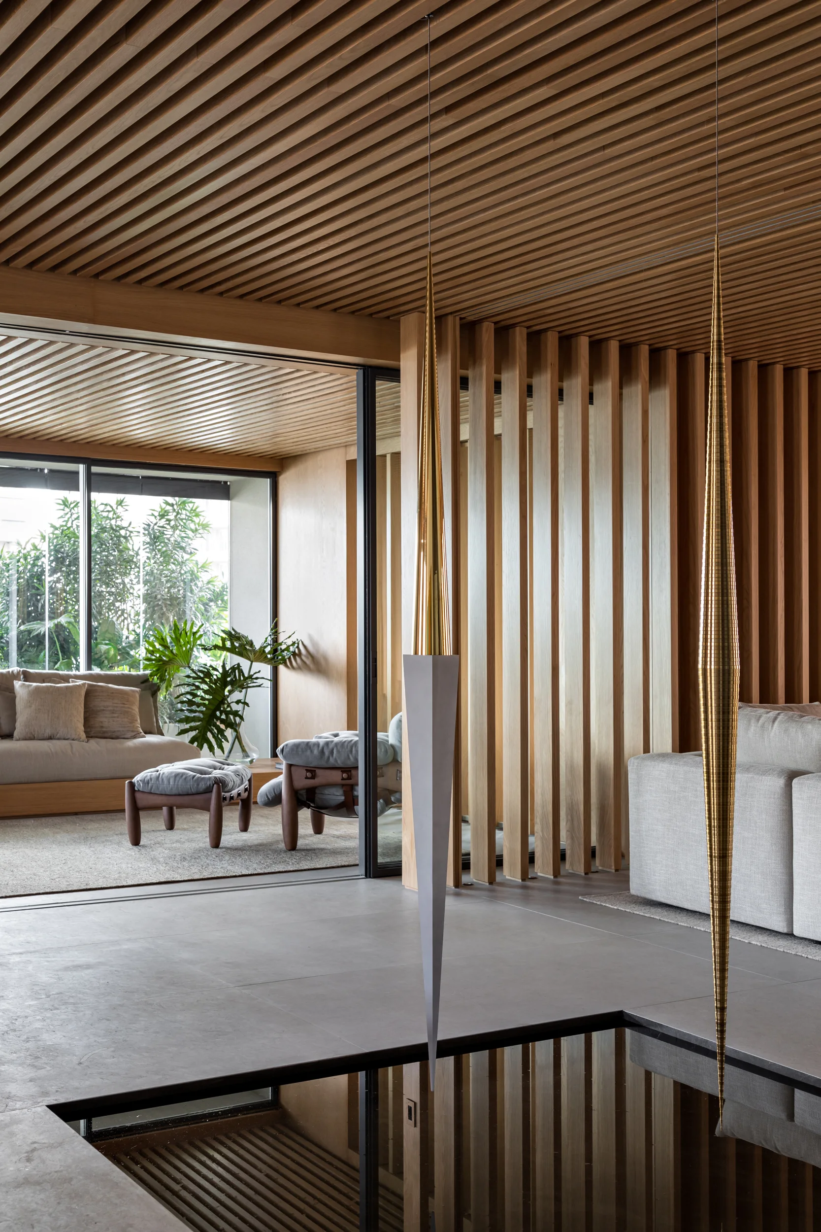A modern living room features a wood slatted ceiling and wall divider, a gray tiled floor, a reflective indoor pool, and sculptural pendant lights.