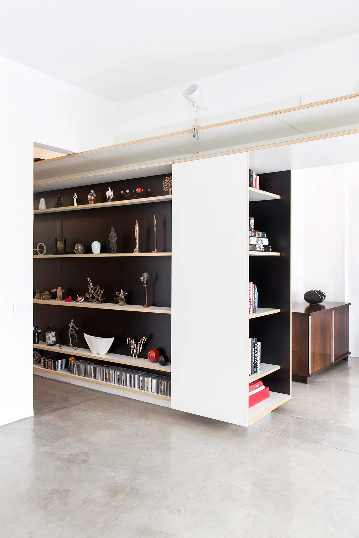 A custom built-in bookshelf with black backing, plywood edges, and a white sliding panel stands on a polished concrete floor.