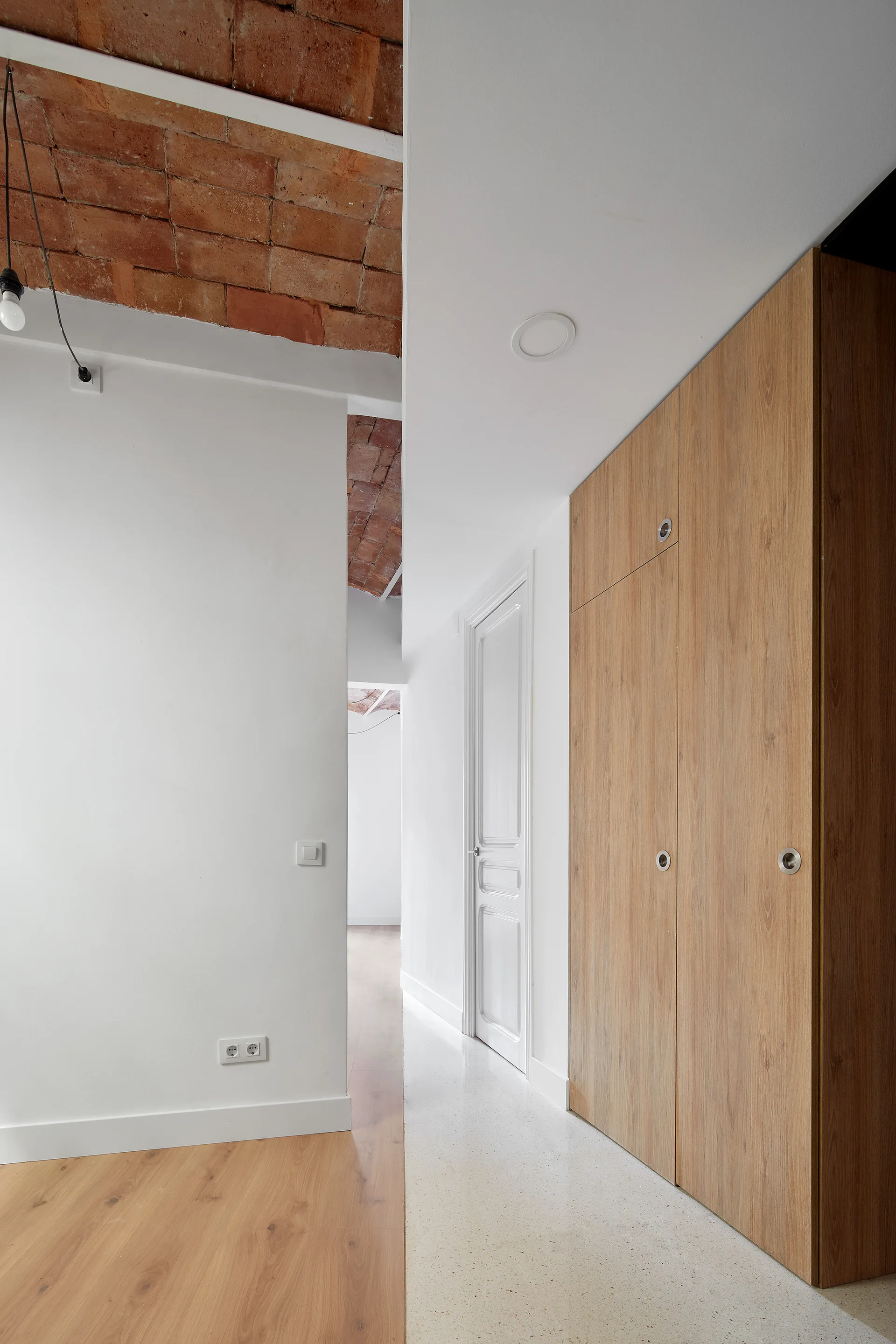 A modern hallway features a vaulted exposed red brick ceiling, plain white walls, a terrazzo-like floor, and tall light-wood built-in wardrobe closets.