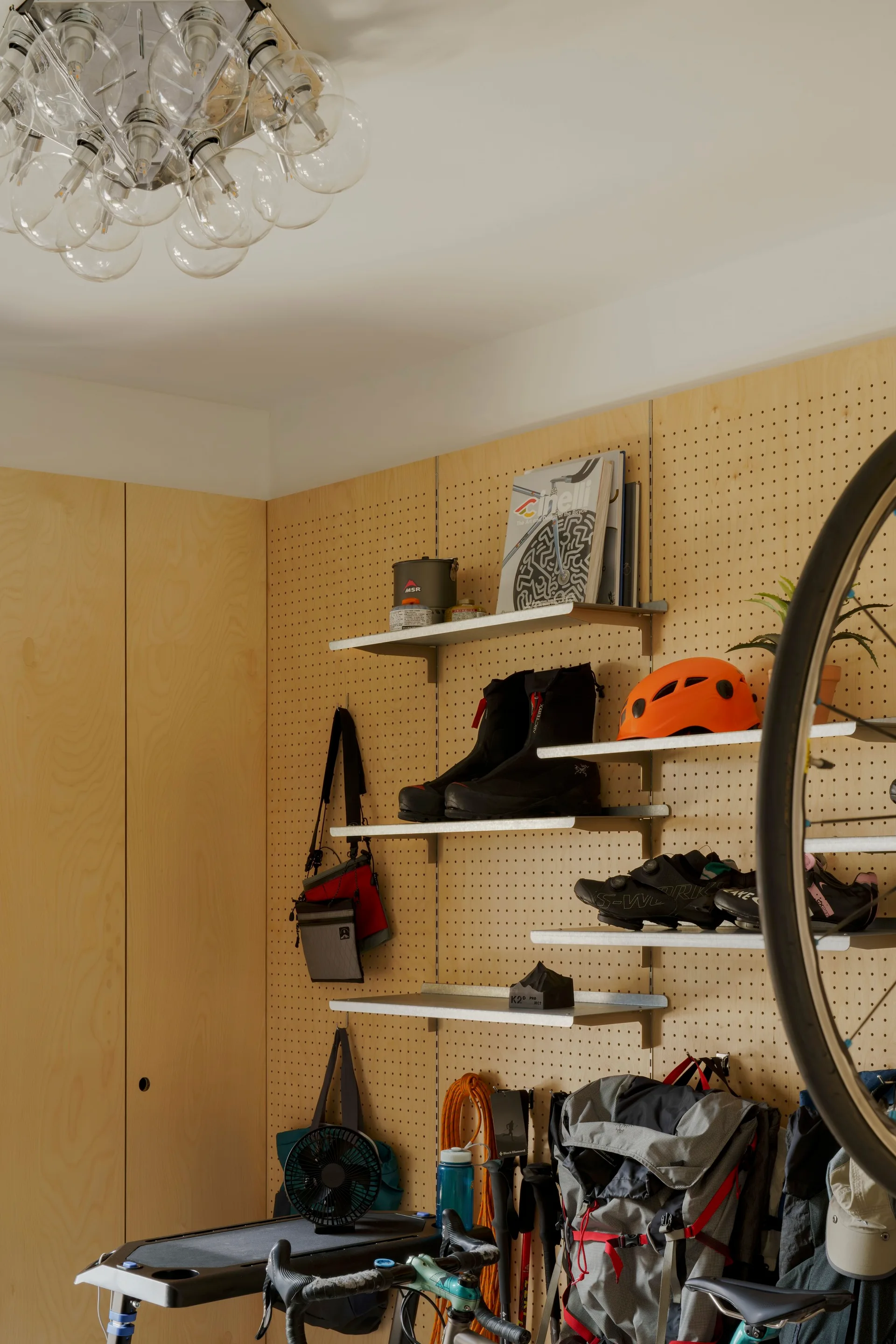 A light wood pegboard wall with metal shelves holding athletic gear, next to a flat panel plywood cabinet and beneath a multi bulb ceiling light.