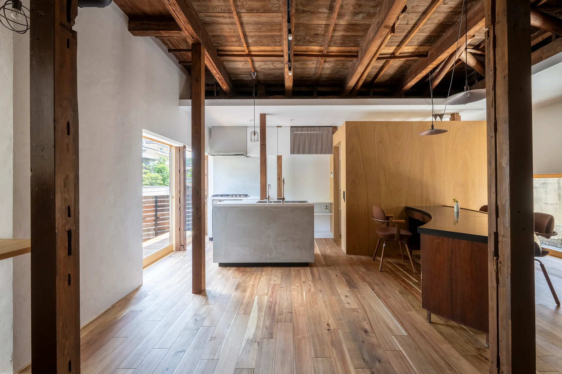 A modern open-plan kitchen featuring exposed timber ceiling beams, a concrete kitchen island, wooden support columns, and a curved wood dining counter.