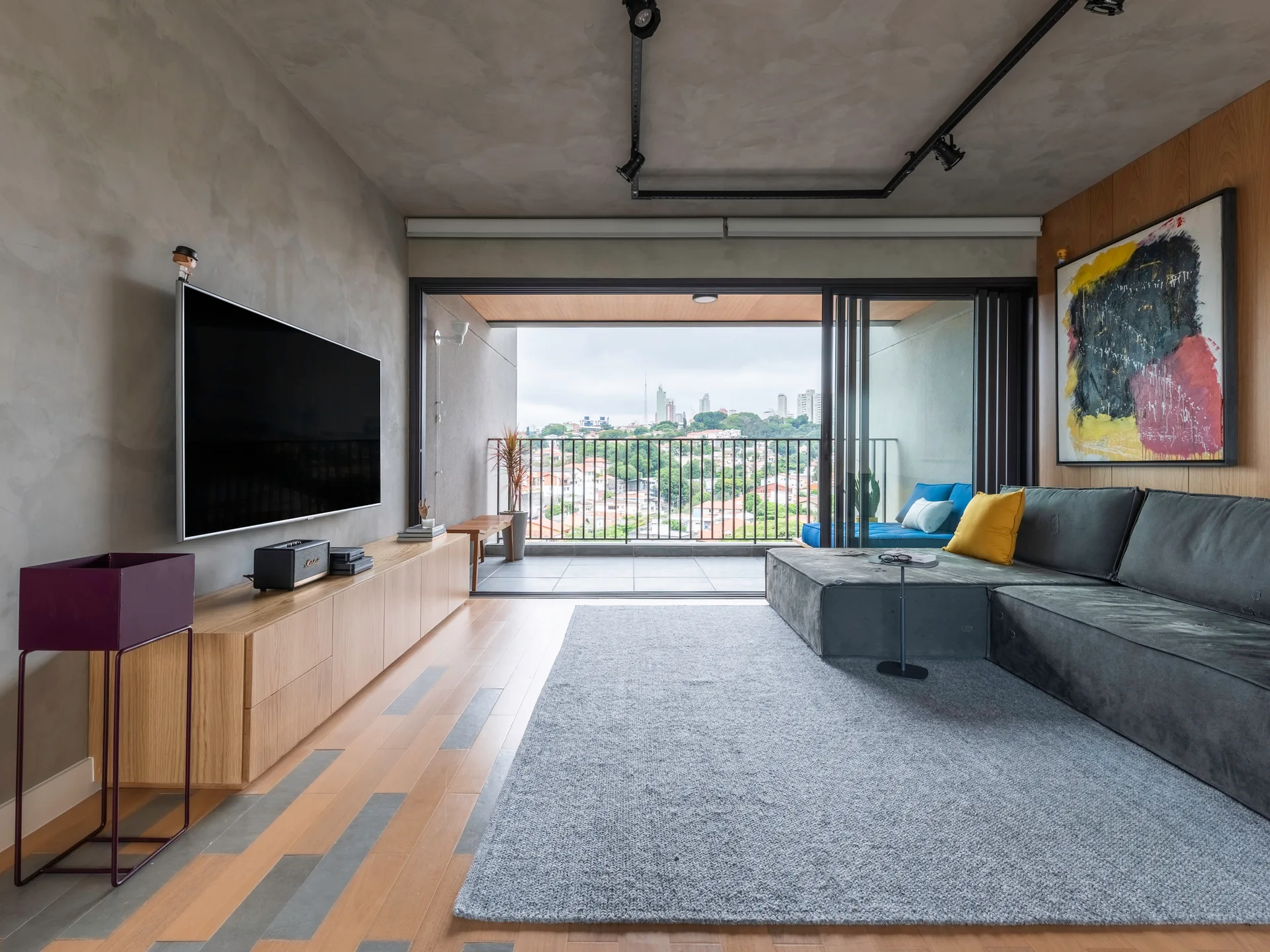 A contemporary living room features a grey sectional sofa, a light wood media console below a television, and sliding glass doors opening to a balcony.