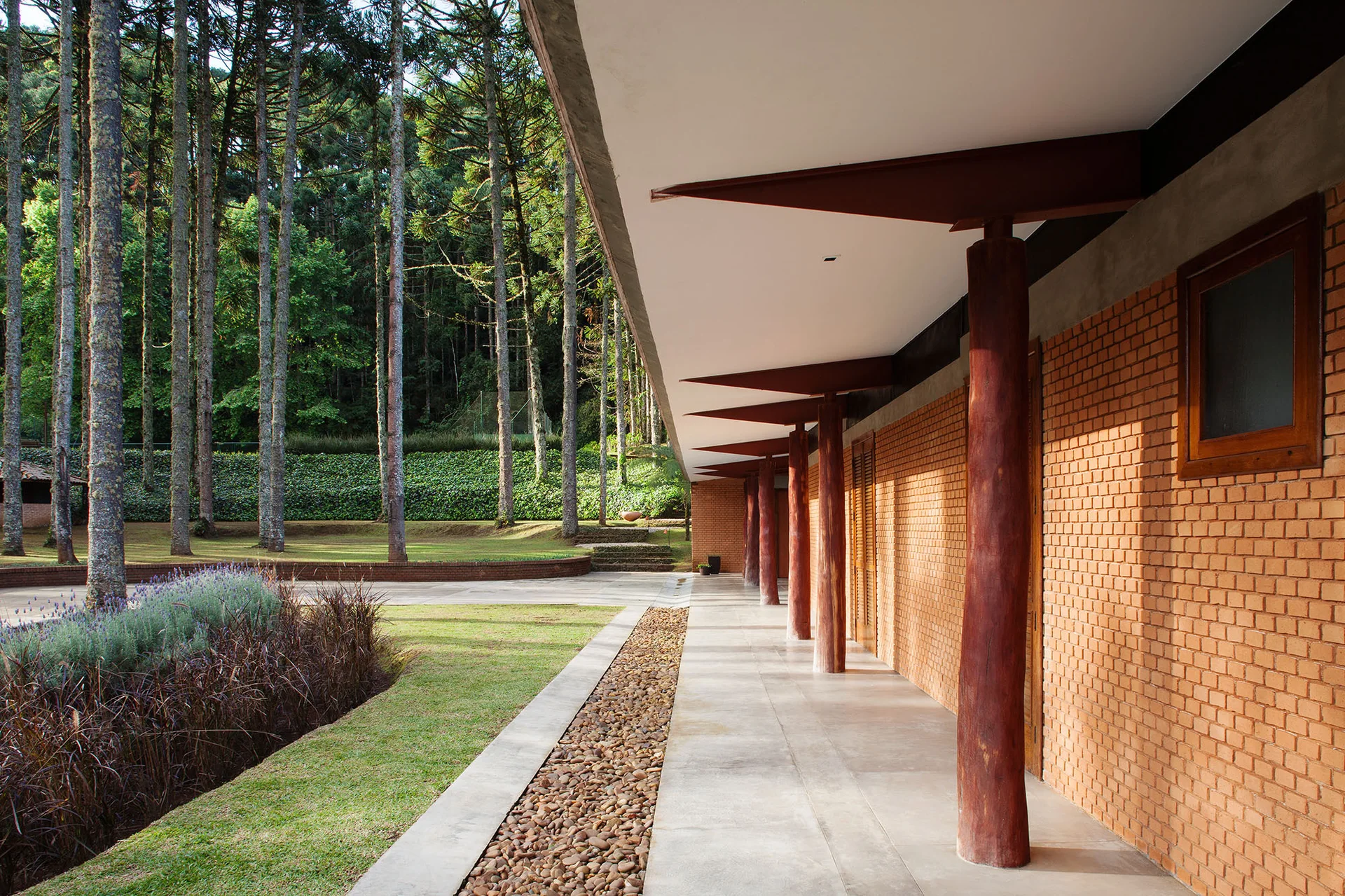 A covered concrete walkway along an orange brick wall features natural log columns supporting a white overhang next to a grassy lawn and pine trees.