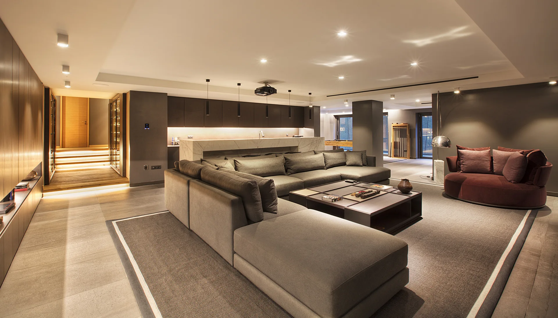 A large grey modular sectional sofa surrounds a dark wood coffee table in a modern open-plan living room with a marble bar and a burgundy armchair.