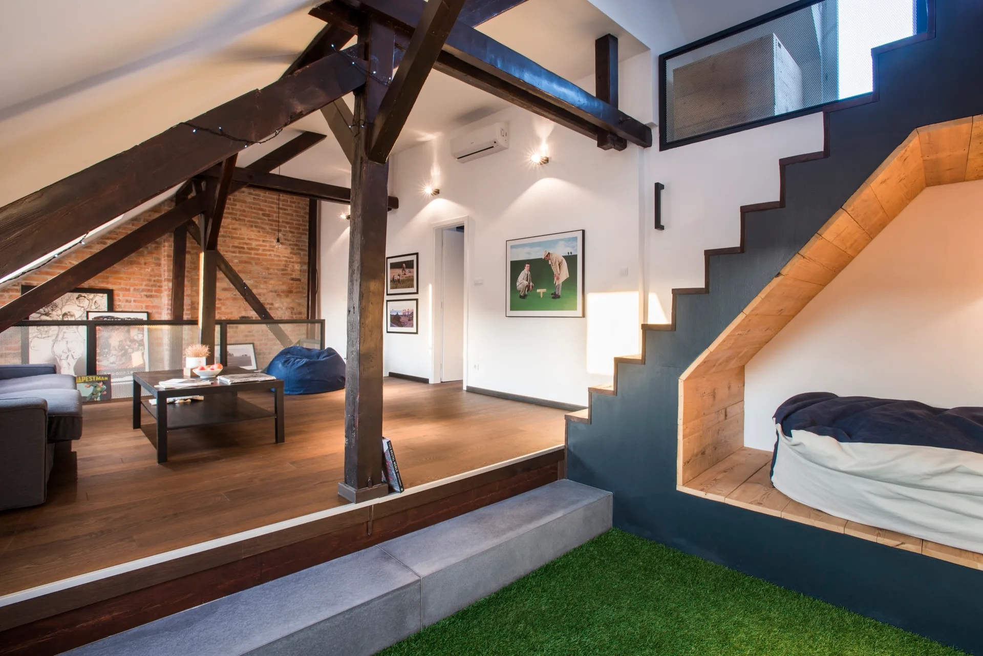 An attic living room with exposed dark wood beams, a brick wall, and a black staircase featuring a built-in wooden reading nook at the bottom.