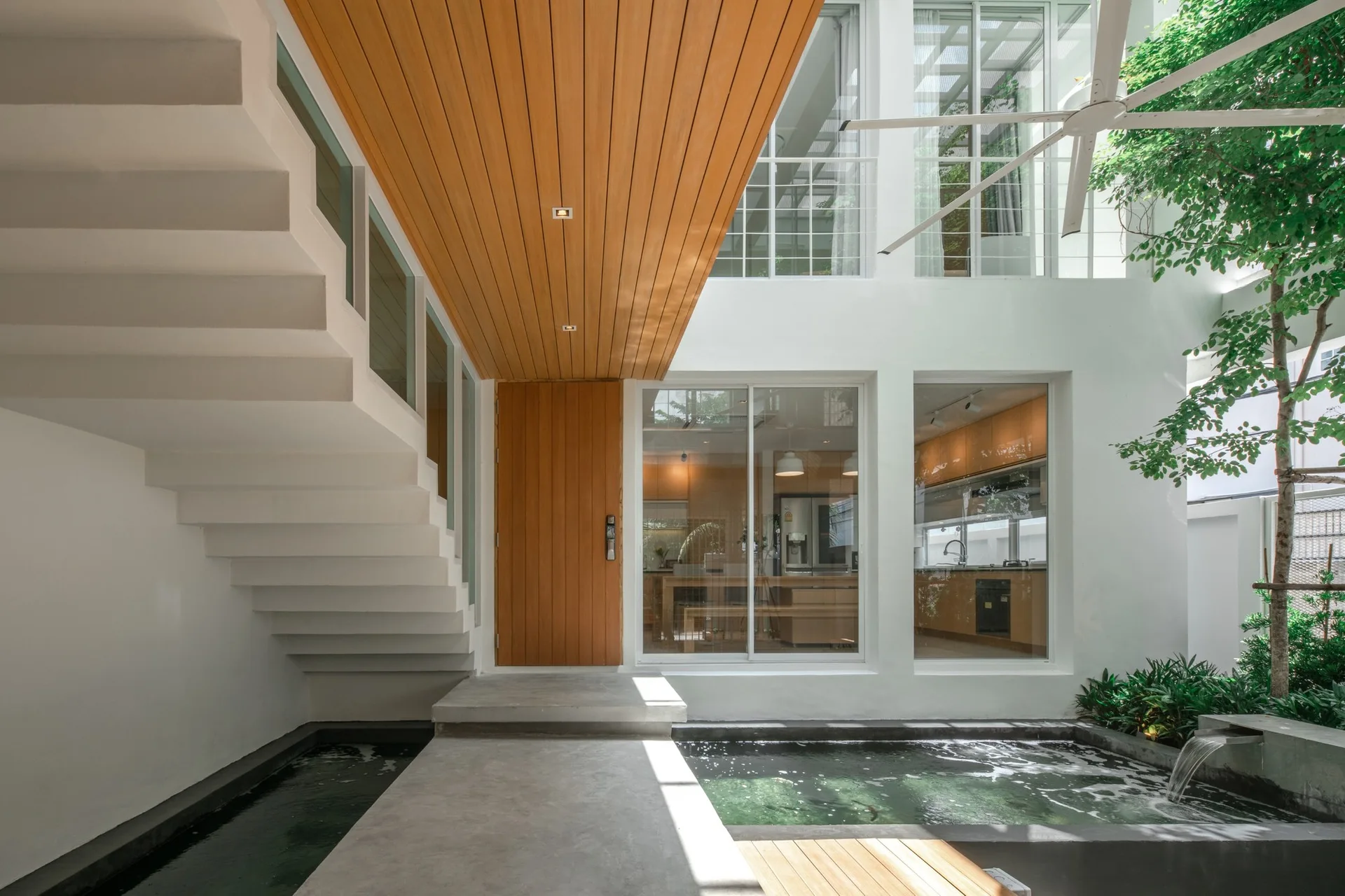 A minimalist entryway features a white floating staircase over an indoor pond, a wood-slatted ceiling, and a concrete walkway leading to a wooden door.
