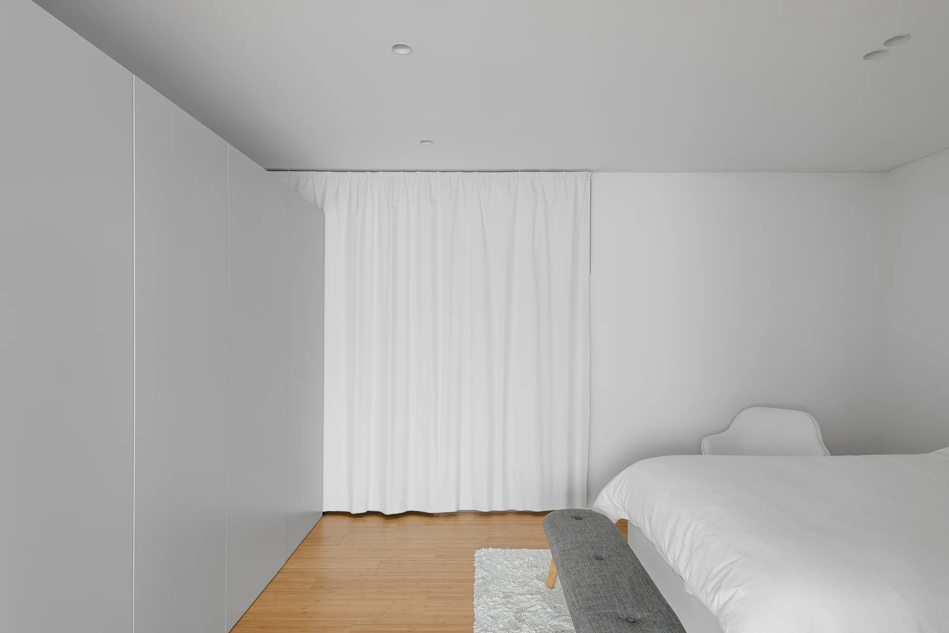 A minimalist bedroom features a large white wardrobe, a bed with white bedding, a gray footstool bench, and light wood floors.