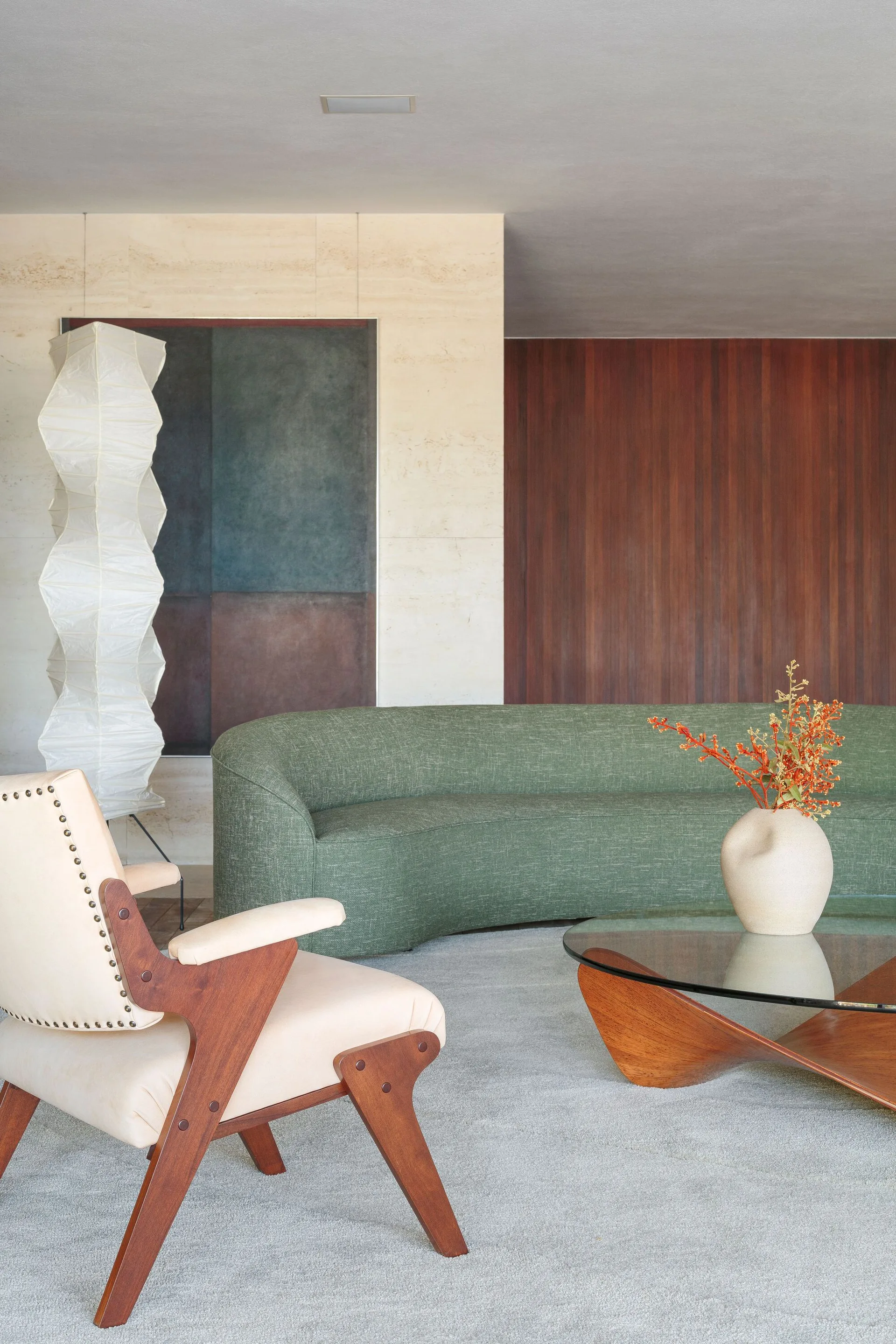 A modern living room features a curved green fabric sofa, a wood-framed armchair, and a glass-topped coffee table against stone and wood-paneled walls.