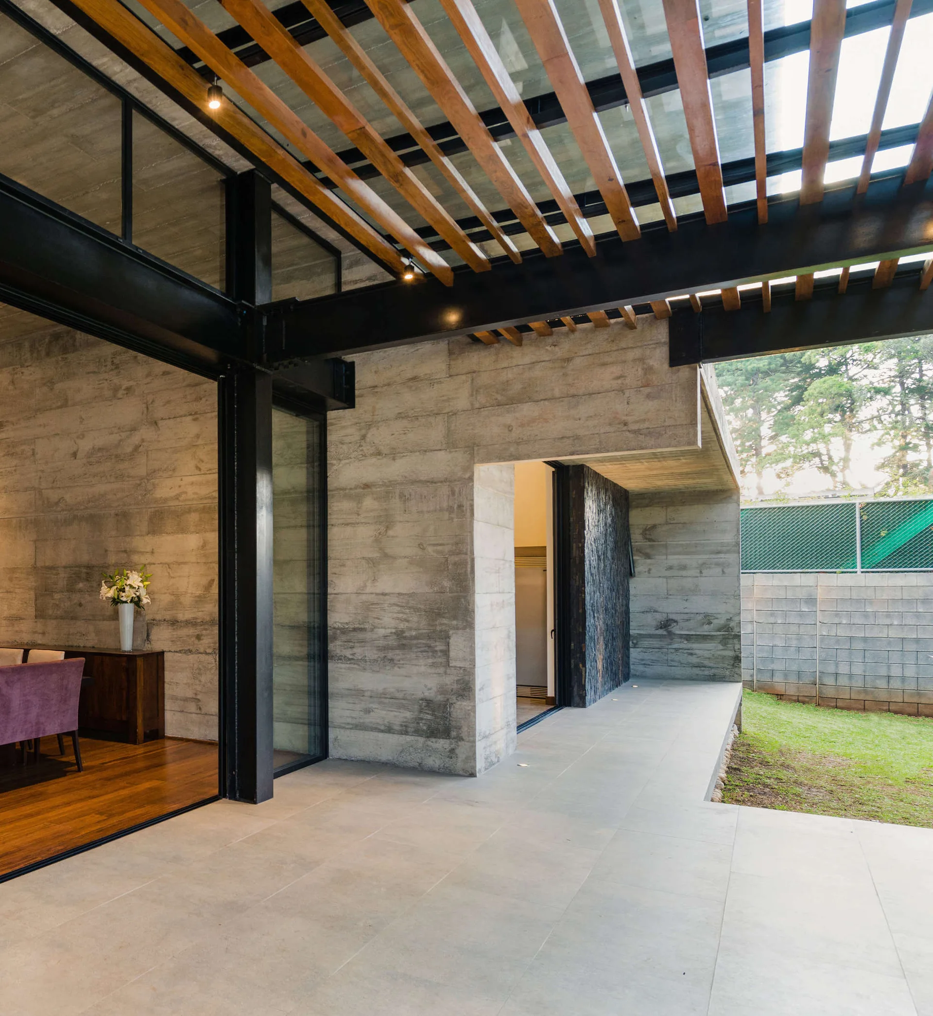 A modern outdoor patio features board-formed concrete walls, black steel structural beams, and a wood-slatted pergola ceiling.