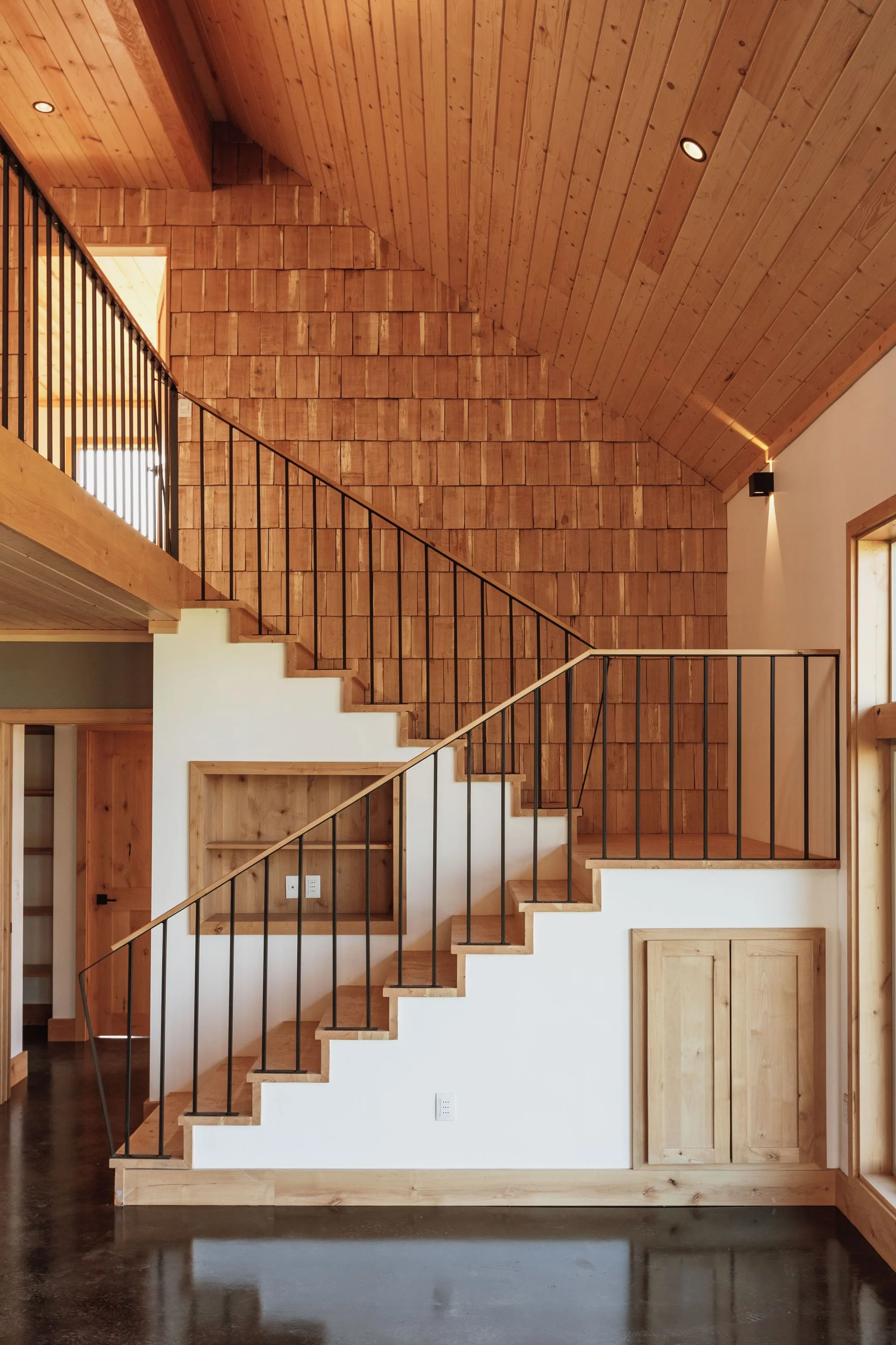 A modern wooden staircase with black metal railings features a wood shingle accent wall, sloped ceiling, and built-in under-stair cabinet.
