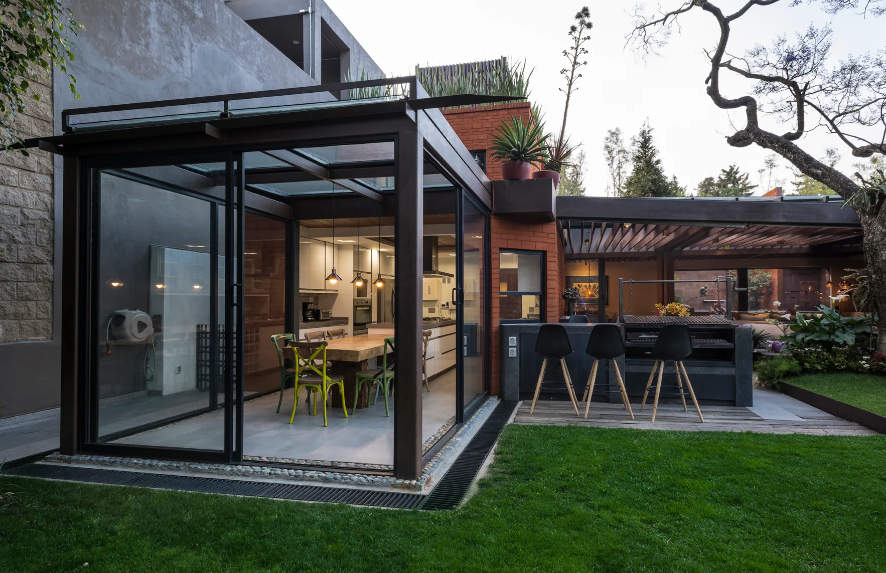 A backyard features a glass-enclosed dining pavilion next to an outdoor kitchen with a stone-topped grill station, black bar stools, and a grassy lawn.