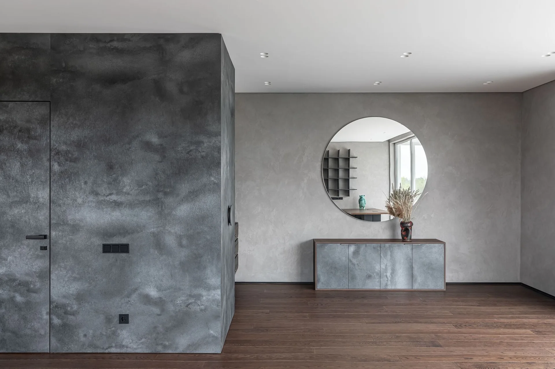 A minimalist entryway features concrete-textured gray walls, a dark wood floor, a low gray console, and a large circular wall mirror.