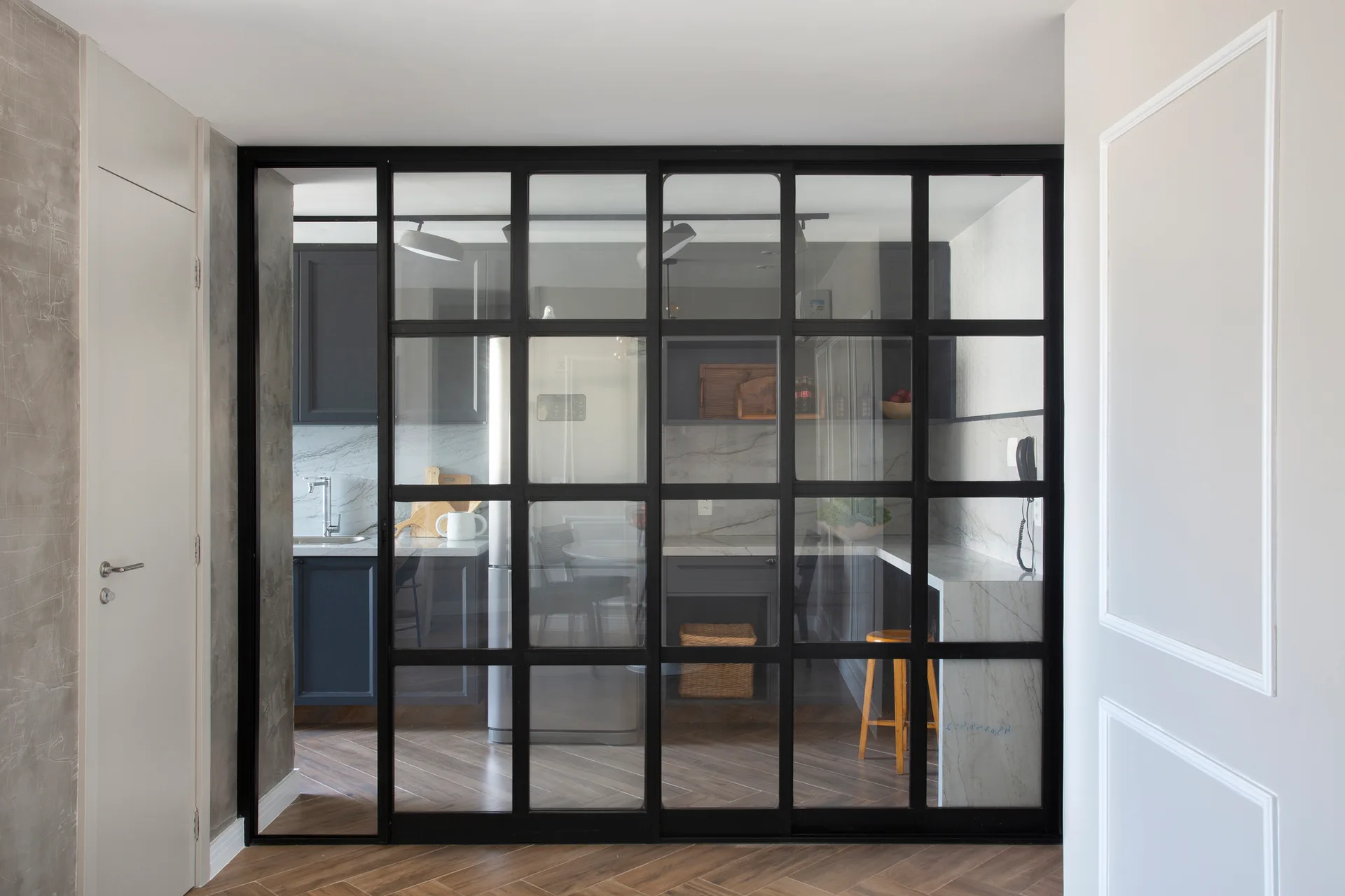 A view through a black-framed glass grid partition wall into a kitchen with dark grey cabinets, marble countertops, and herringbone flooring.