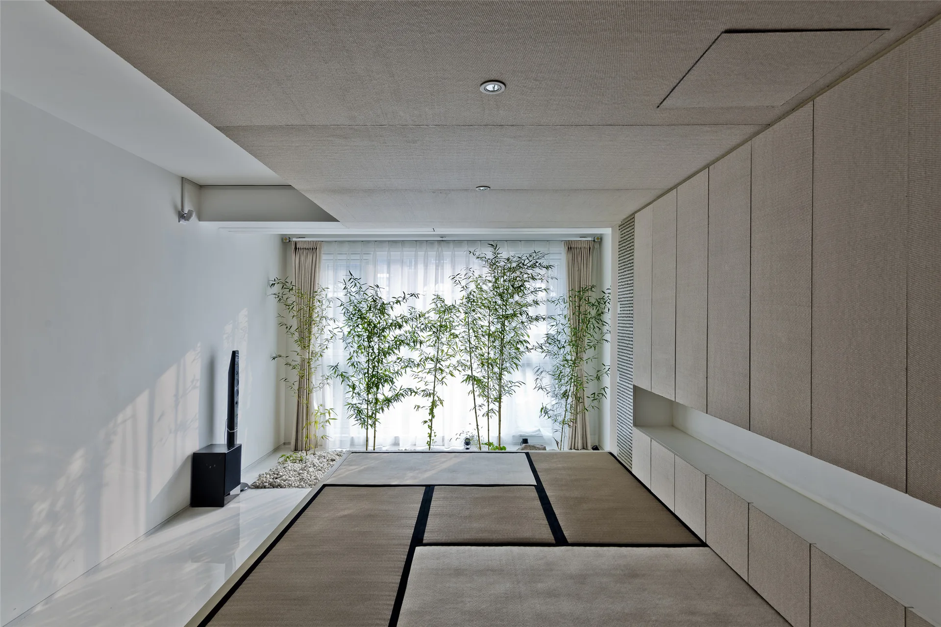 A minimalist room featuring tatami mats on the floor, fabric-textured built-in cabinetry, and bamboo plants growing near a sunlit window.