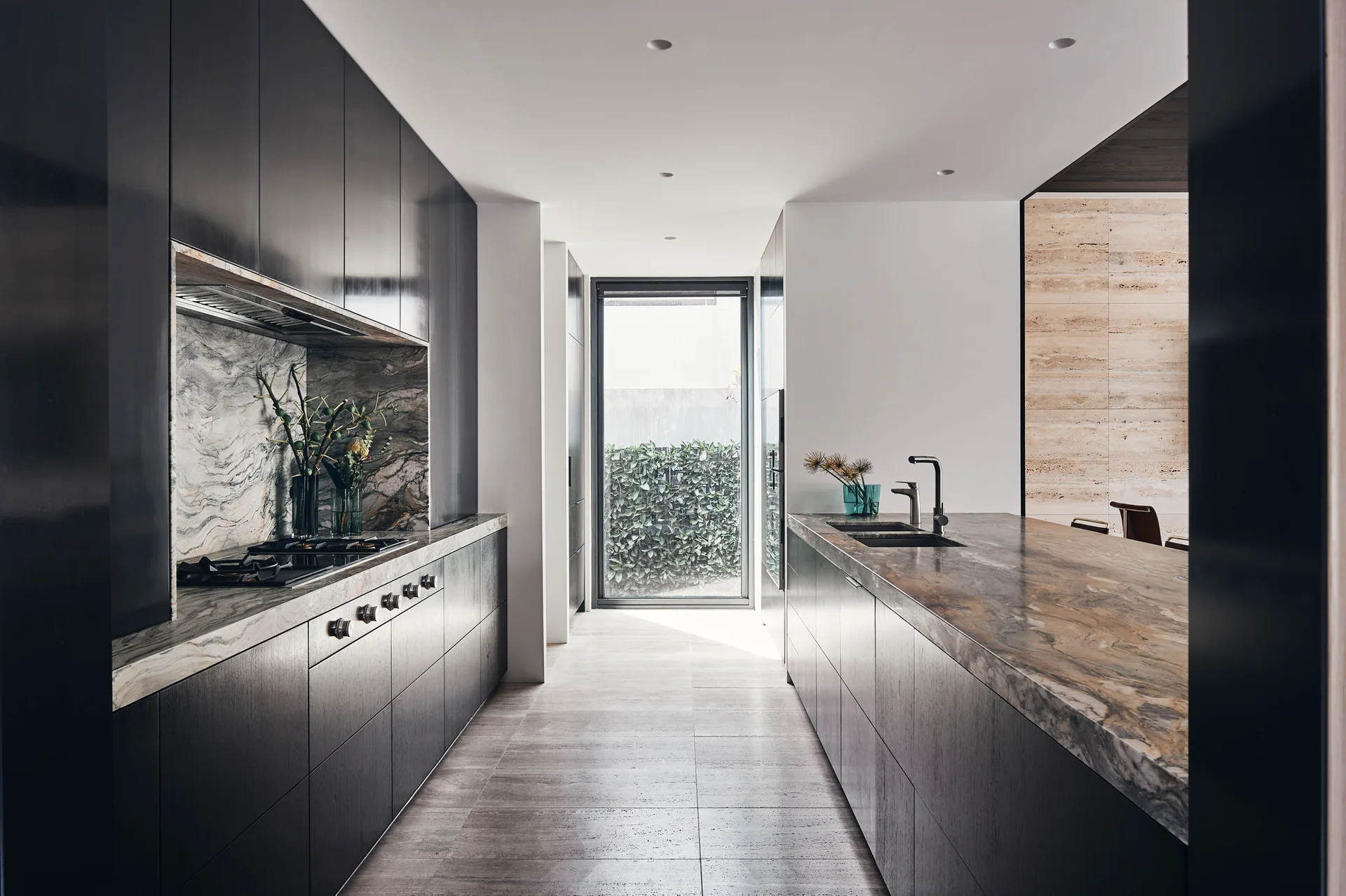 A modern galley kitchen featuring dark charcoal cabinetry, a deeply veined stone countertop, a matching slab backsplash, and a window displaying a hedge.