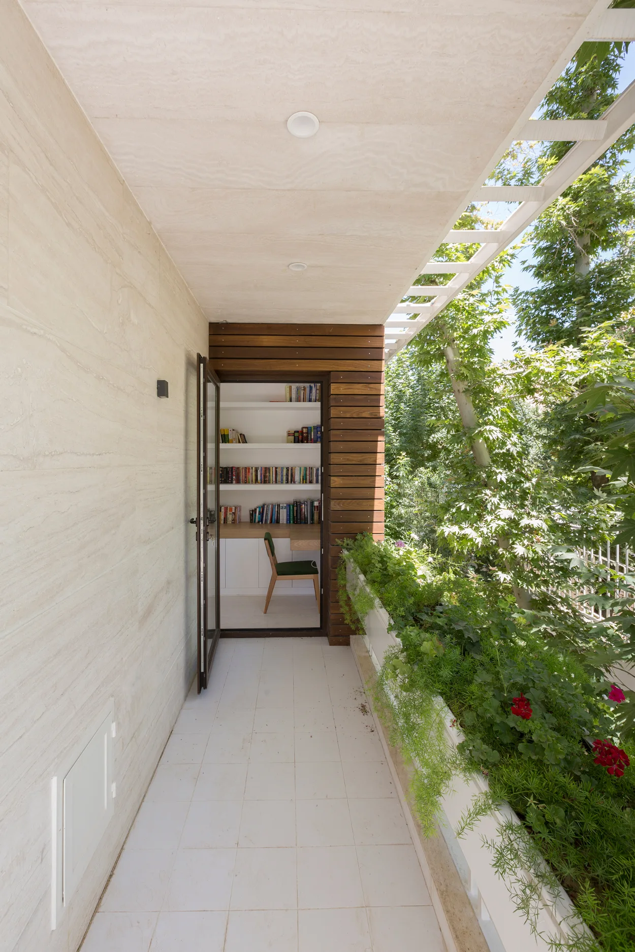 A tiled outdoor balcony with a green planter box leads through a glass door into a small home library with built-in bookshelves.