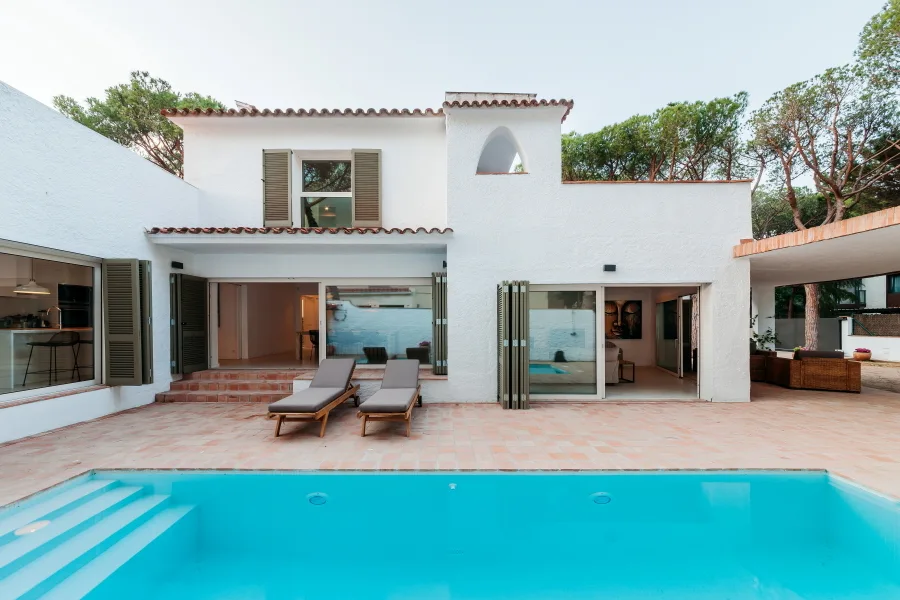 A white stucco Mediterranean house with olive green folding shutters faces a blue swimming pool and a terracotta tile patio with two lounge chairs.