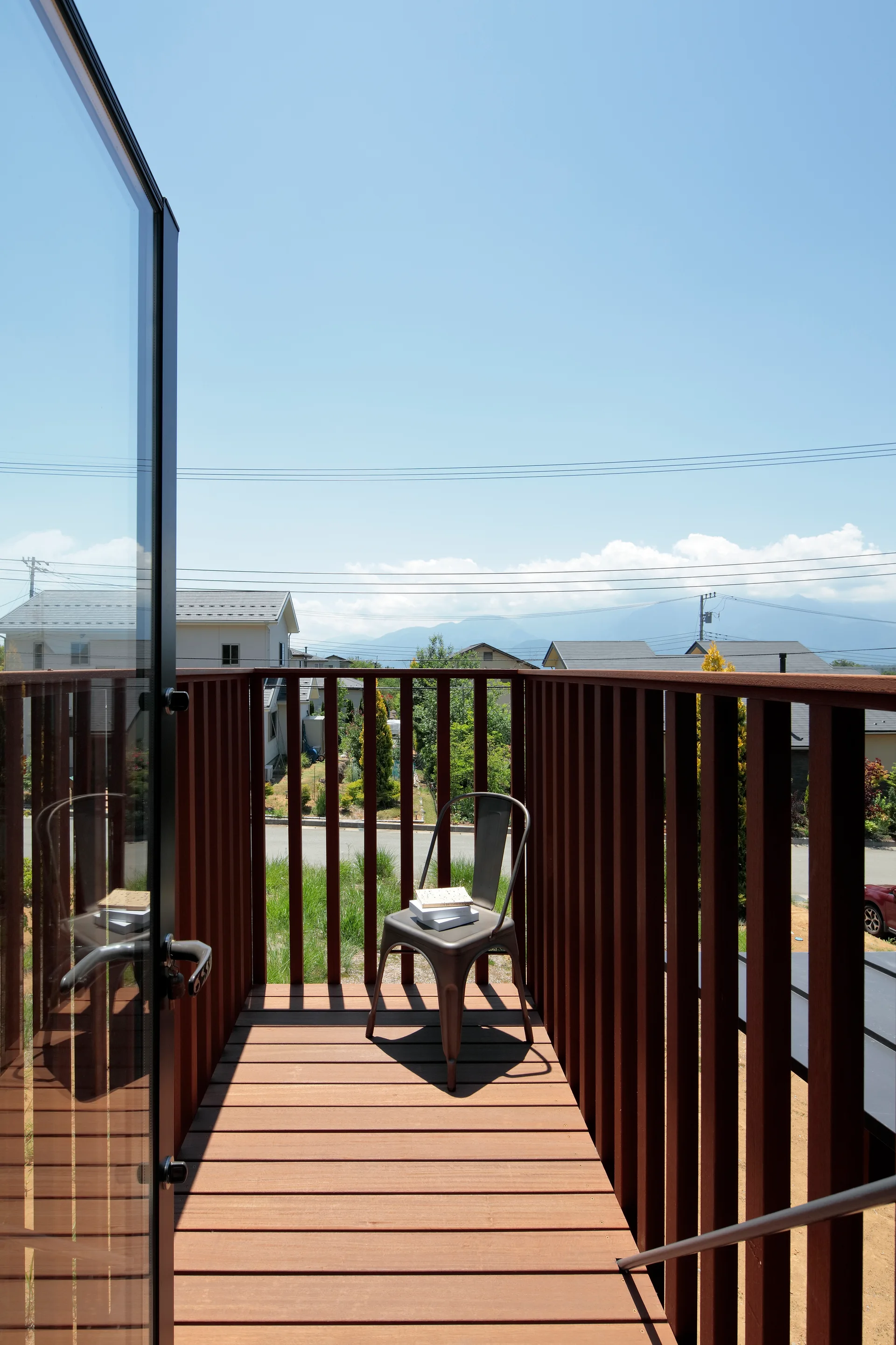 A narrow wooden balcony with vertical slatted railings holds a single metal chair under a clear blue sky.
