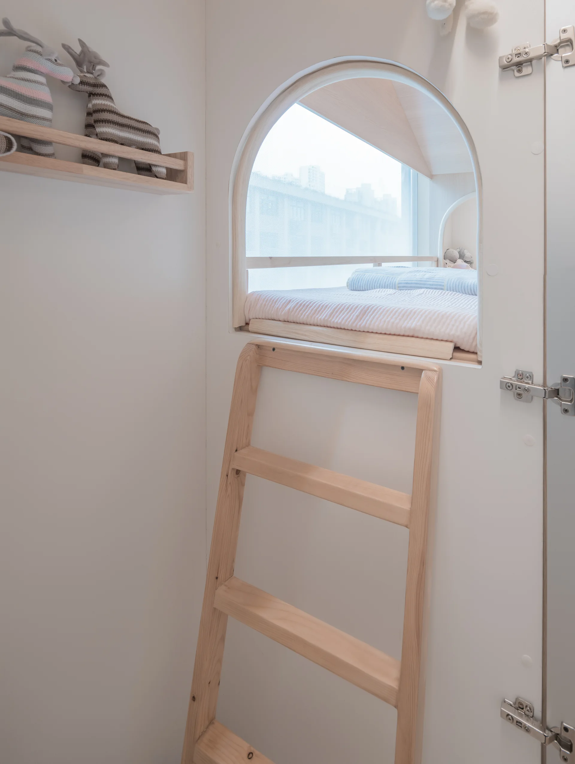 A white built-in loft bed nook features an arched opening, a light wood ladder, and a matching wall shelf holding stuffed toys.