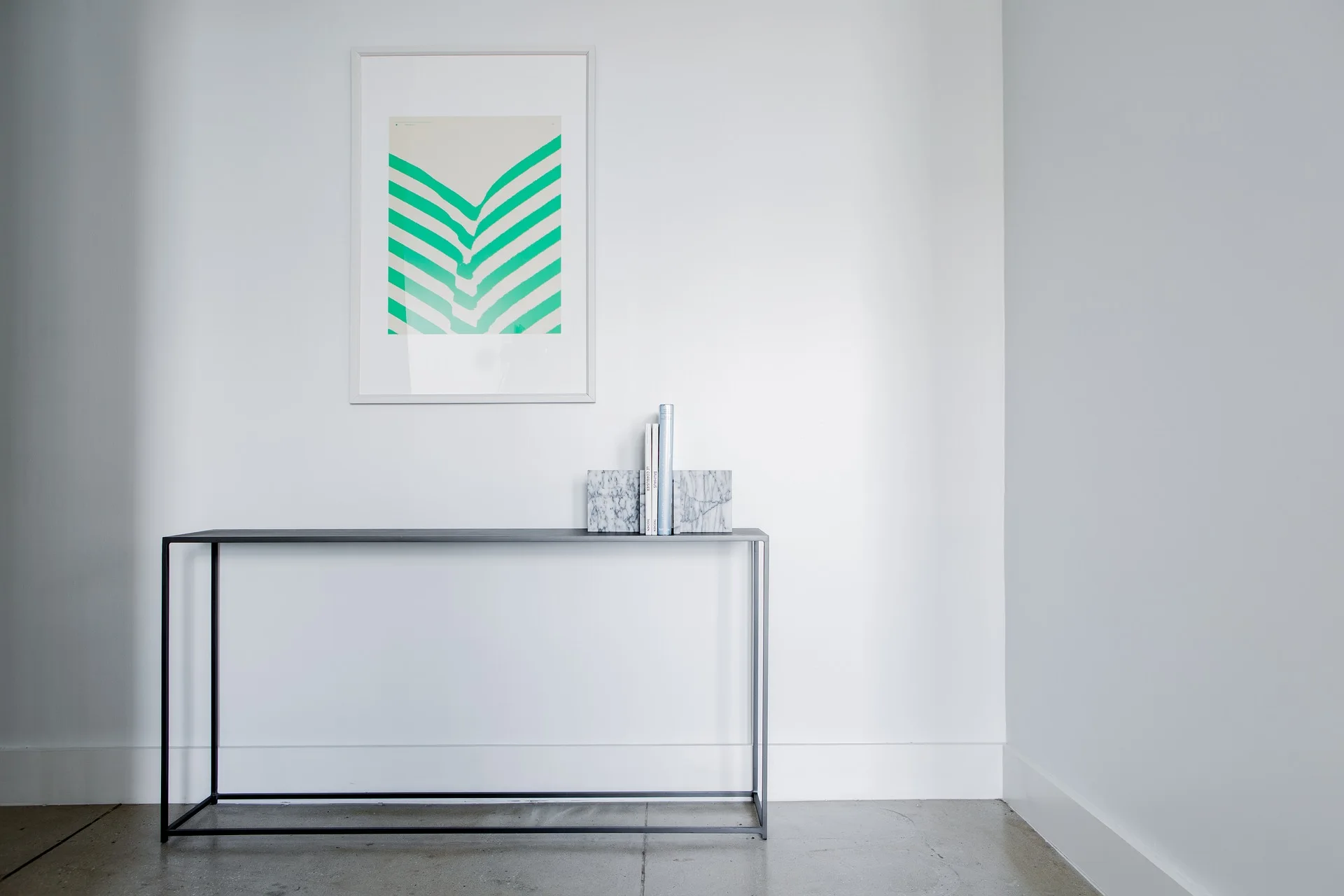 A slim black metal console table sits against a plain white wall next to marble bookends and a framed green graphic print.