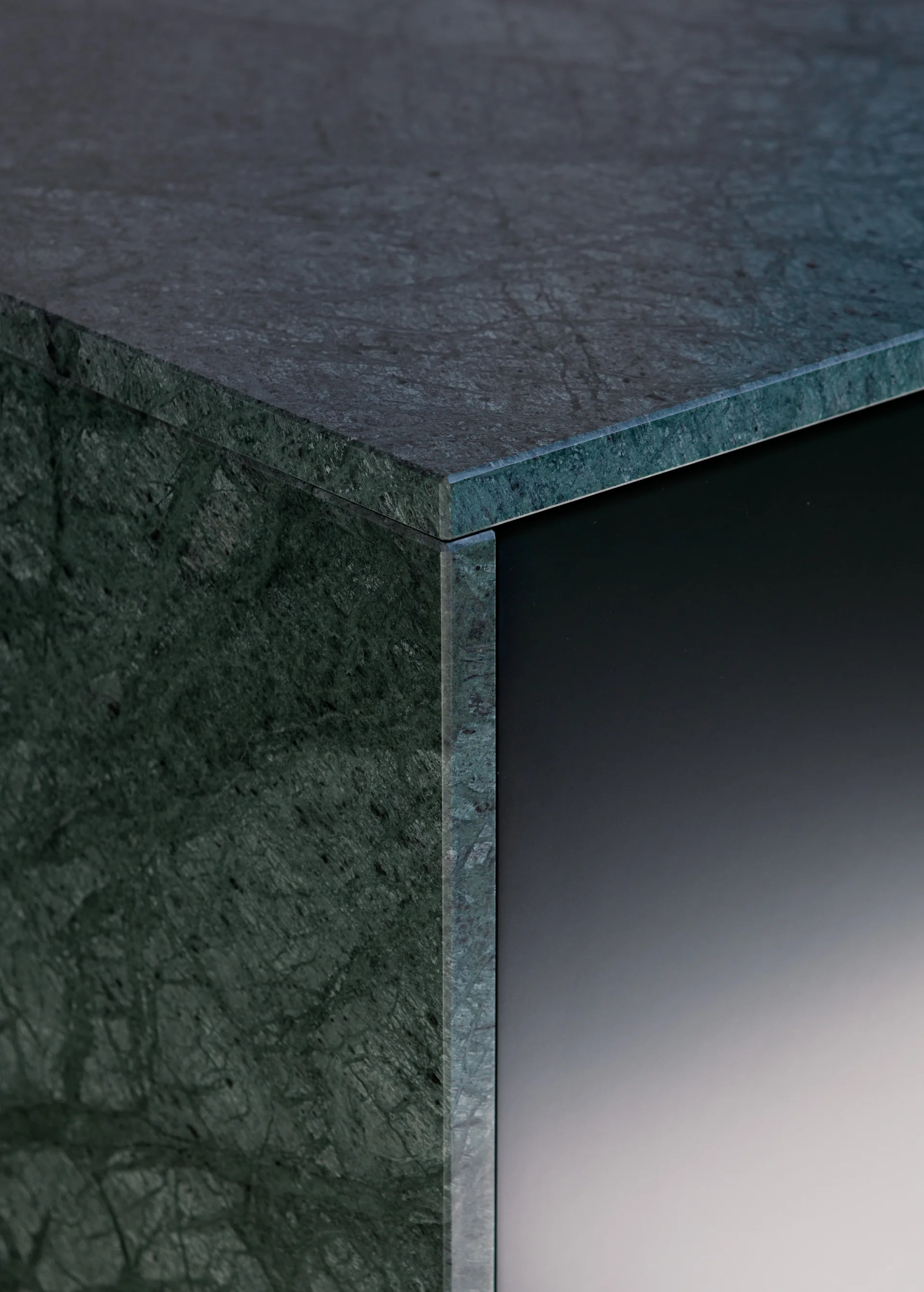 A close-up view of a dark green veined marble countertop corner with a sharp mitered edge next to a smooth matte black cabinet panel.