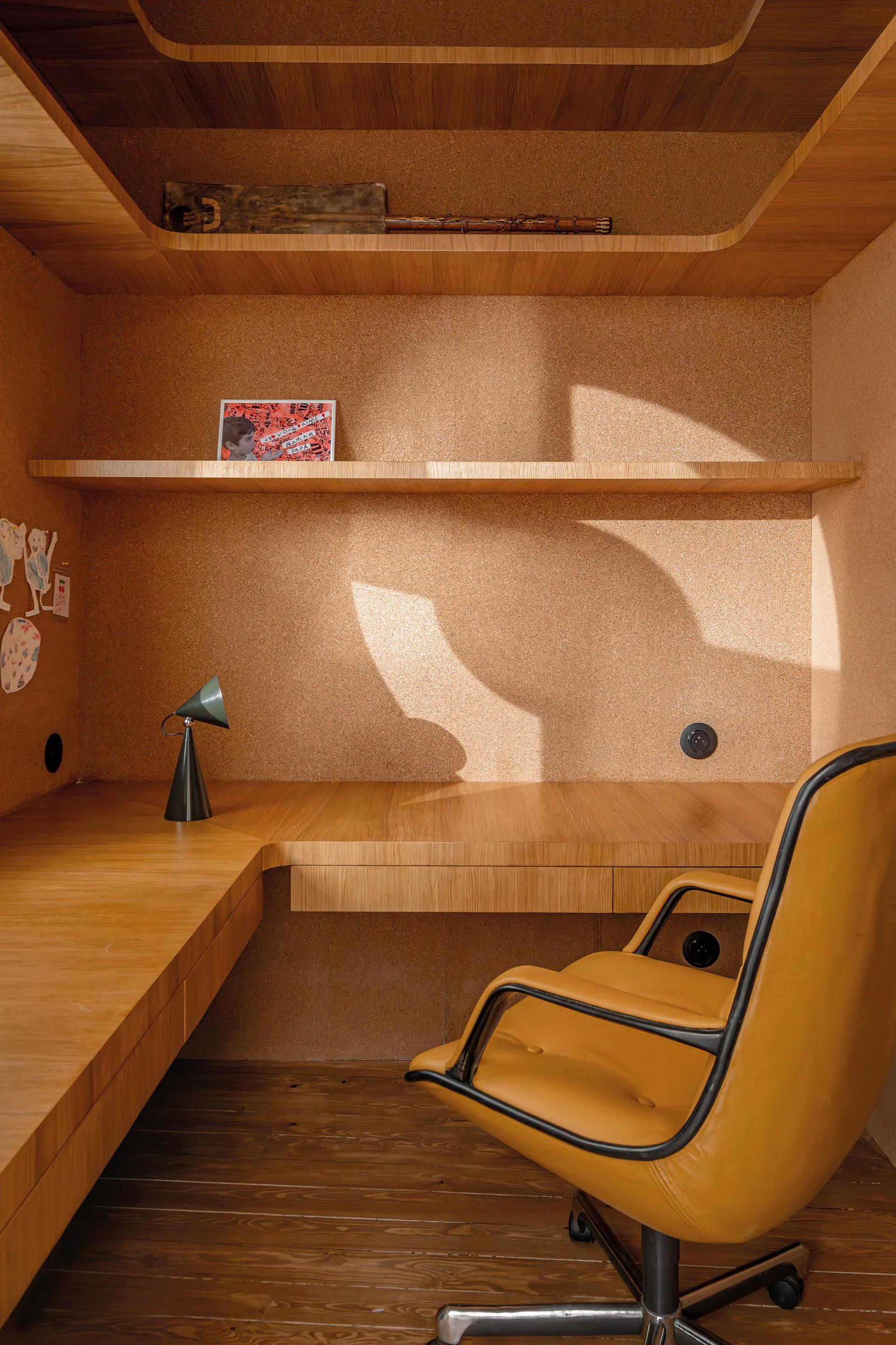 A built-in wooden desk niche features textured cork walls, a floating wood shelf, a green task lamp, and a mustard yellow leather office chair.
