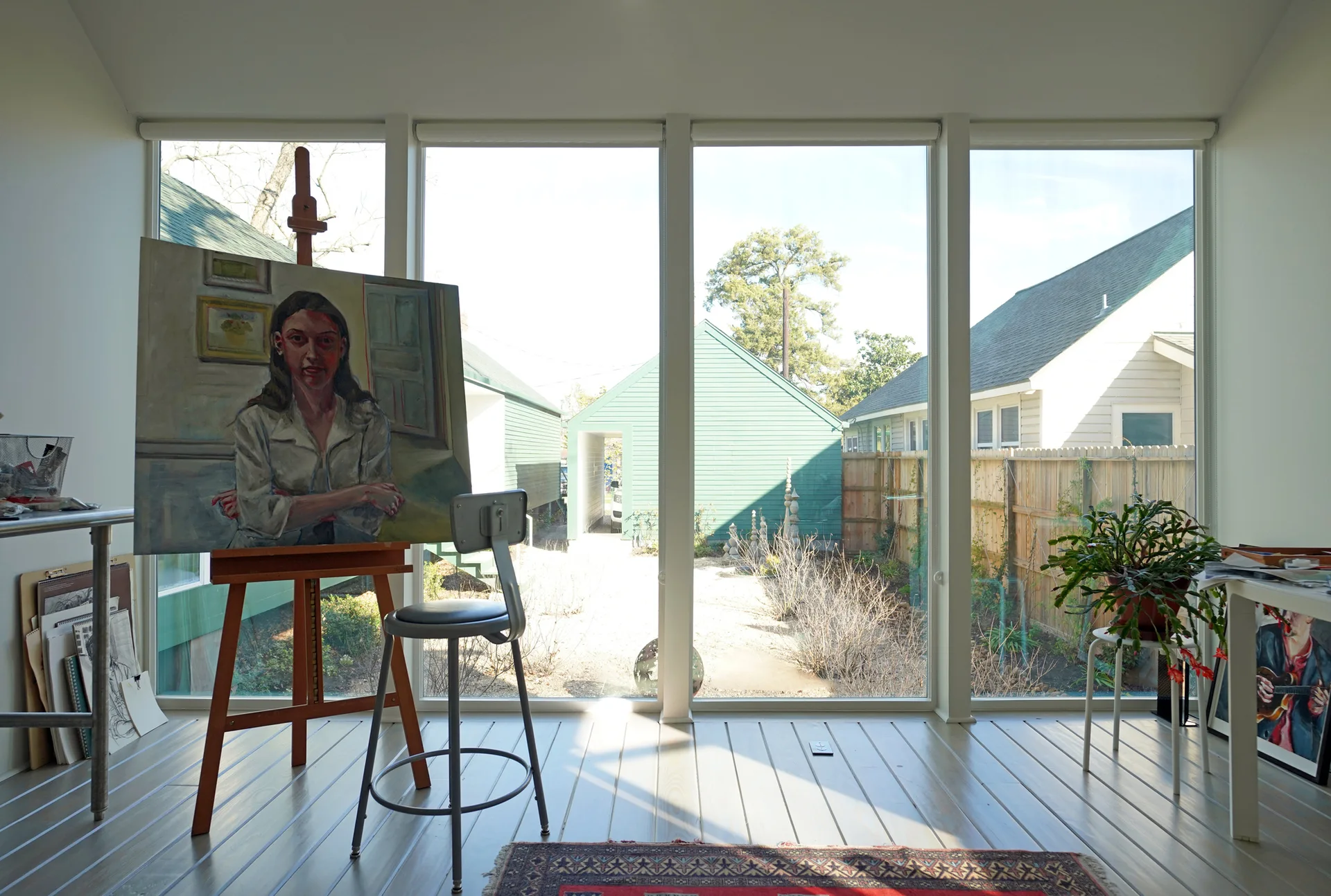 A bright art studio features a wooden easel with a portrait painting, a grey metal stool, and large windows looking out onto a green backyard.