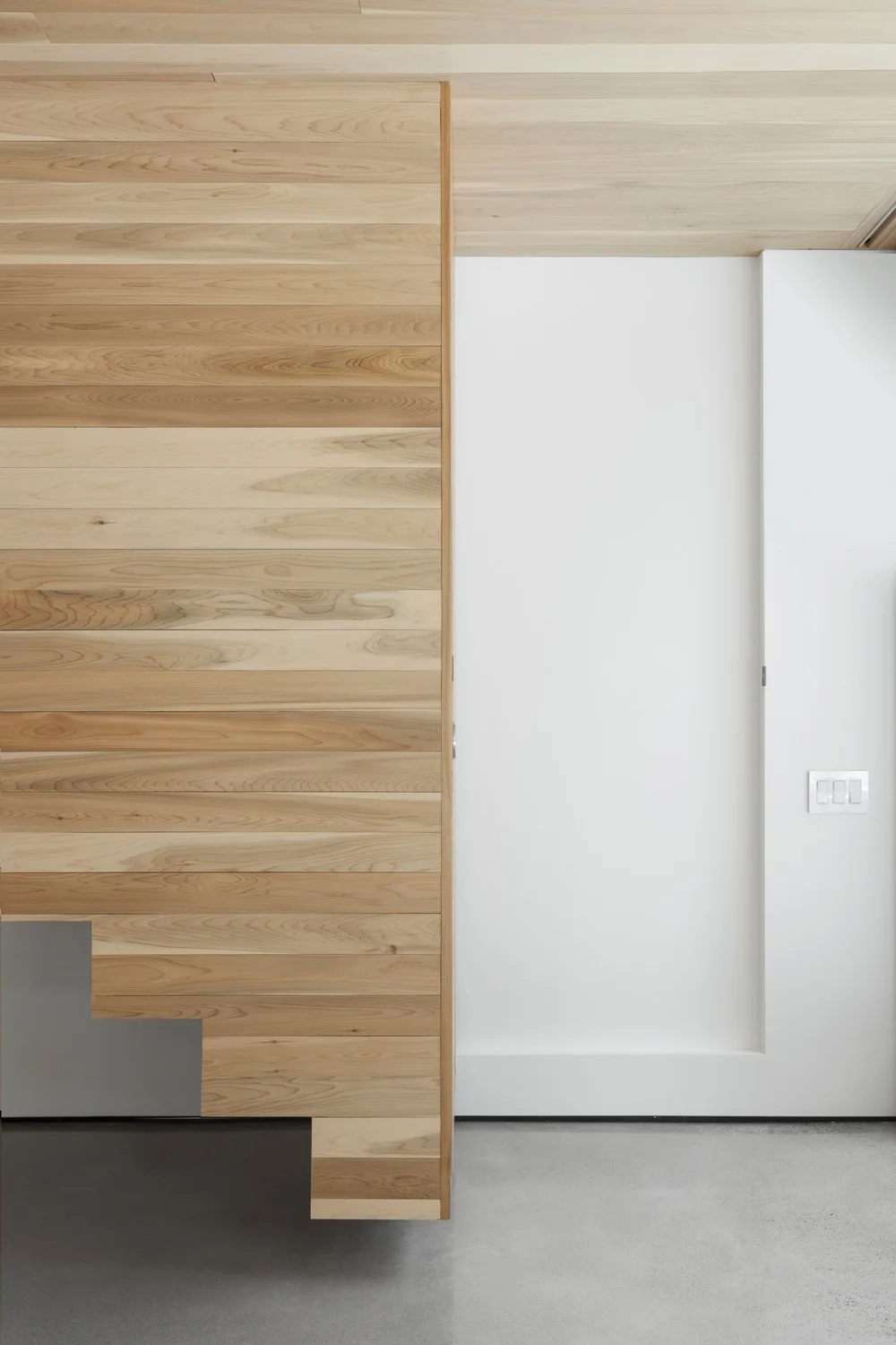 An interior view of horizontal light wood cladding meeting a smooth white wall and door above a grey concrete floor.