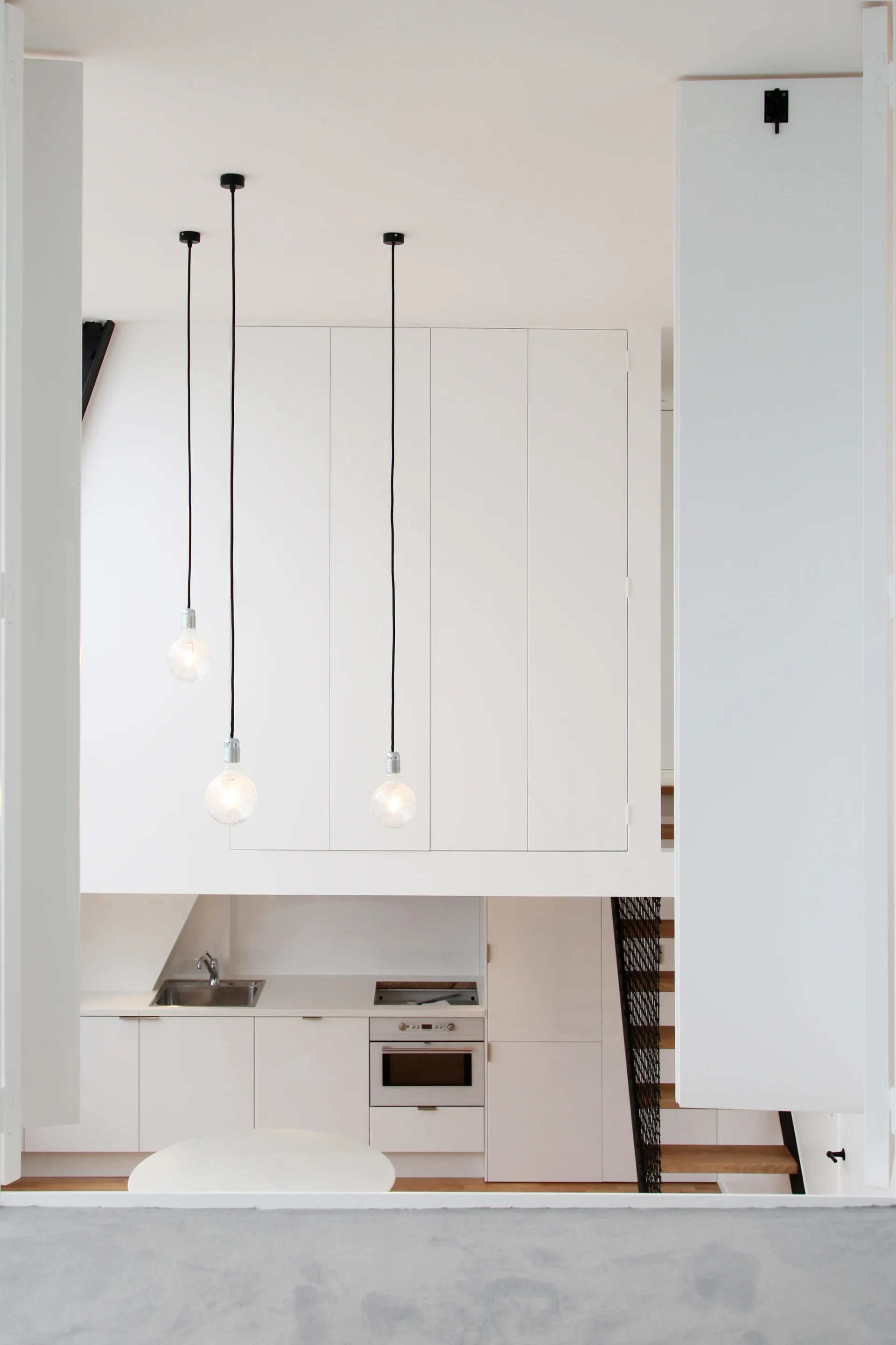 A minimalist white kitchen features flat-front cabinets, three hanging globe pendant lights, and a wooden staircase in the background.